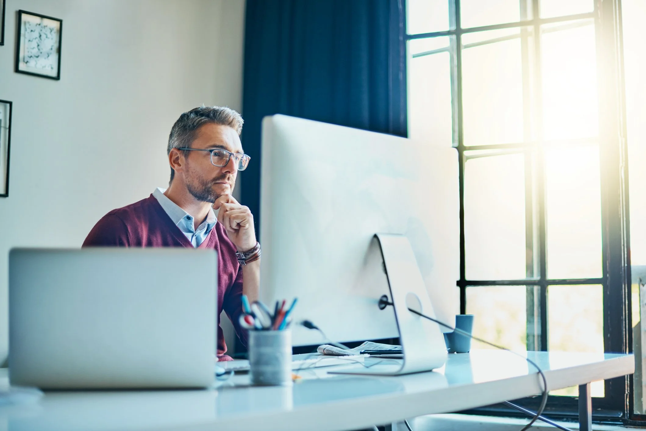 Man with glasses working at a desk in front of a computer, looking thoughtful in a bright office with large windows.