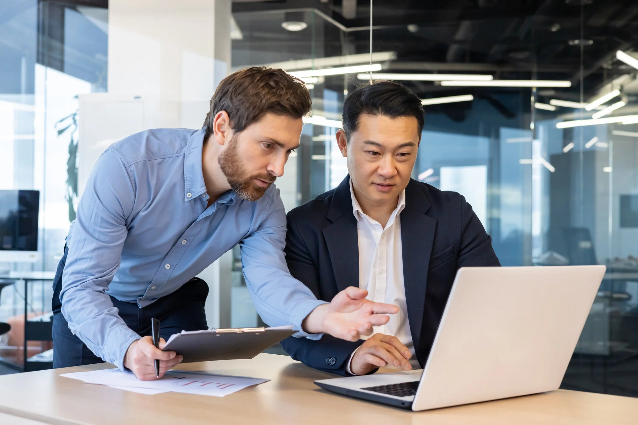 Two men working together in an office, looking at a laptop screen. One man is seated, and the other is leaning over, pointing at the laptop. They appear to be discussing work.
