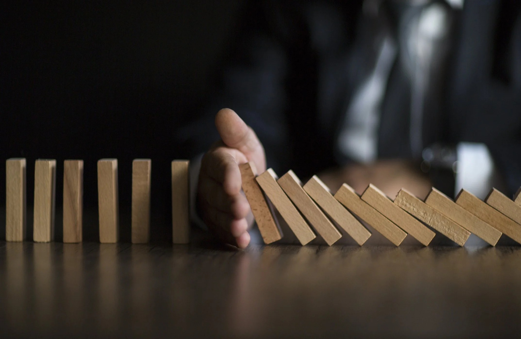 A hand stopping a line of falling wooden dominoes from toppling over on a dark surface.