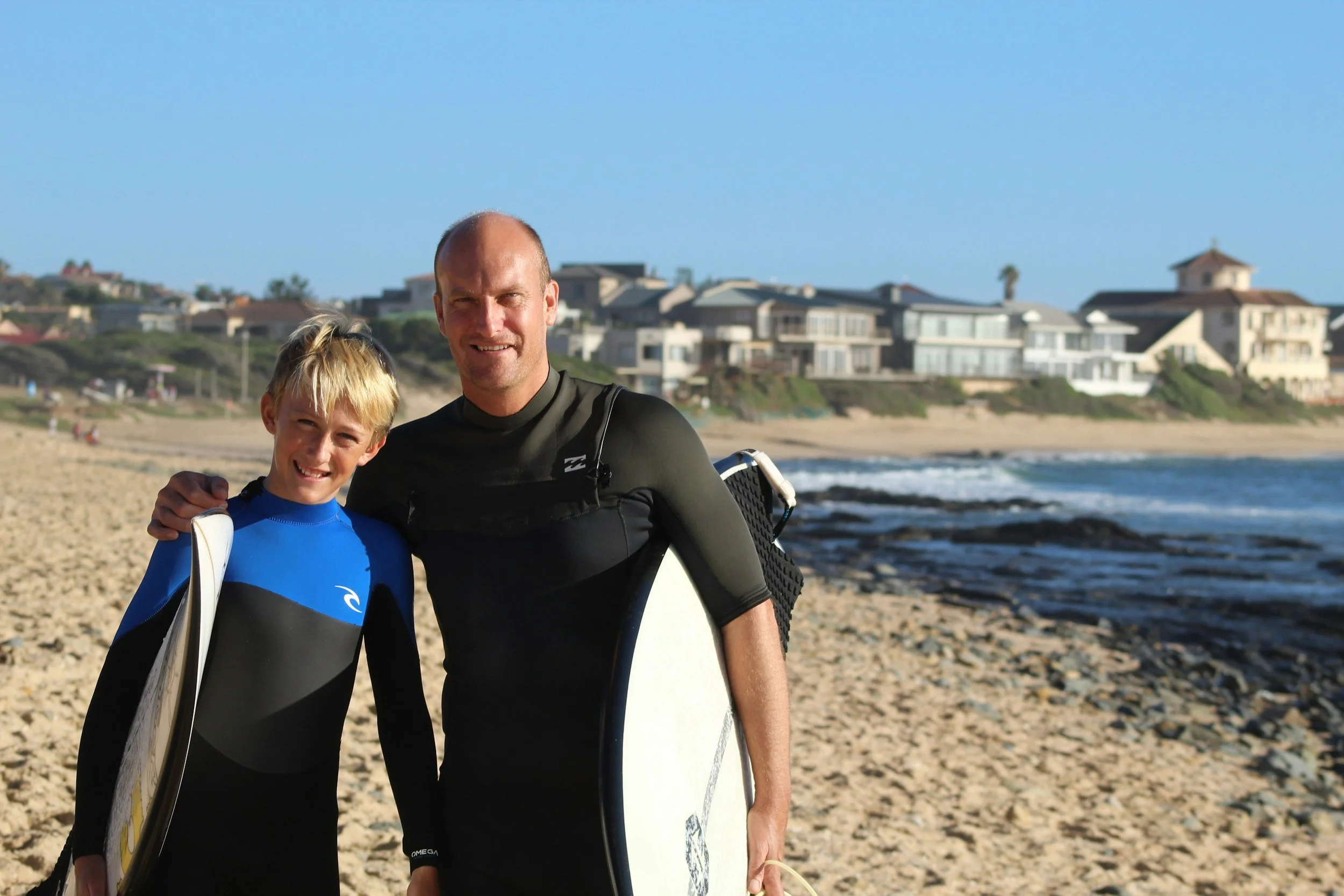 A father and son in wetsuits holding surfboards on a beach with houses in the background.