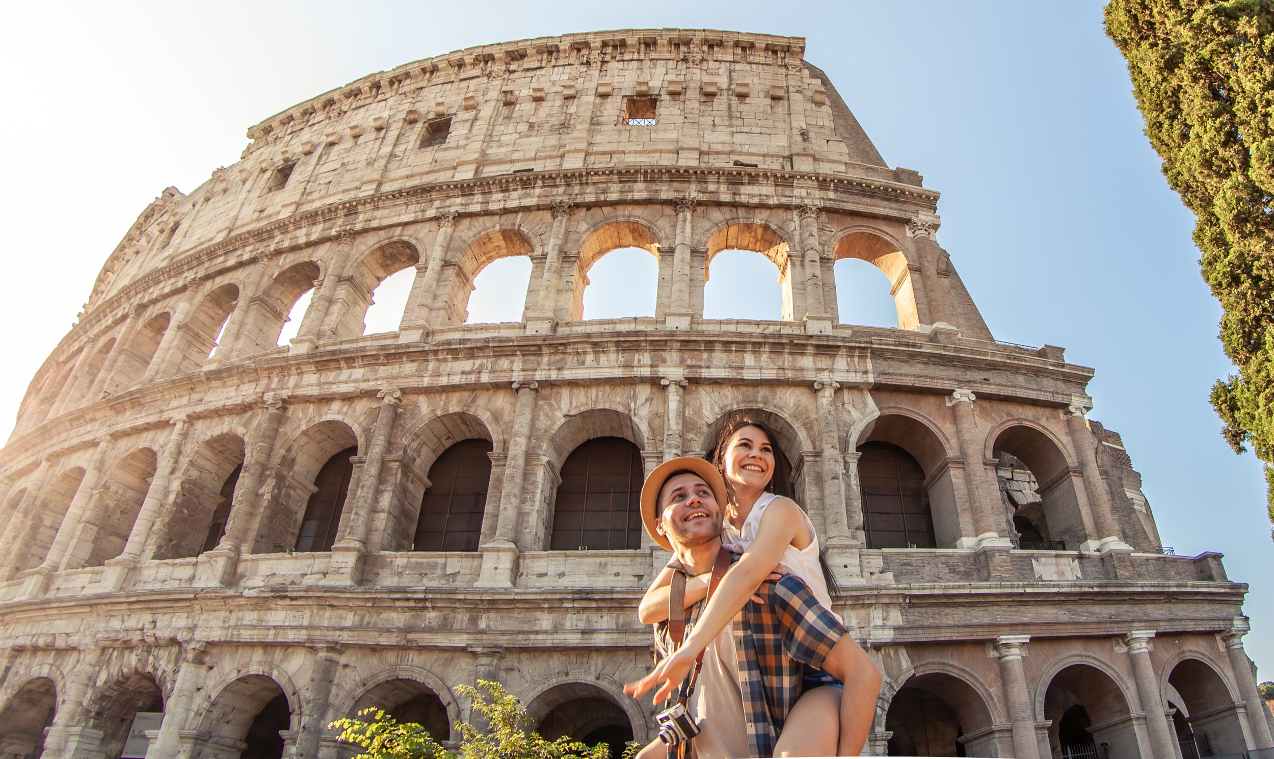 Smiling couple, a man carrying a woman, in front of the Colosseum in Rome, Italy during daytime with clear blue sky.