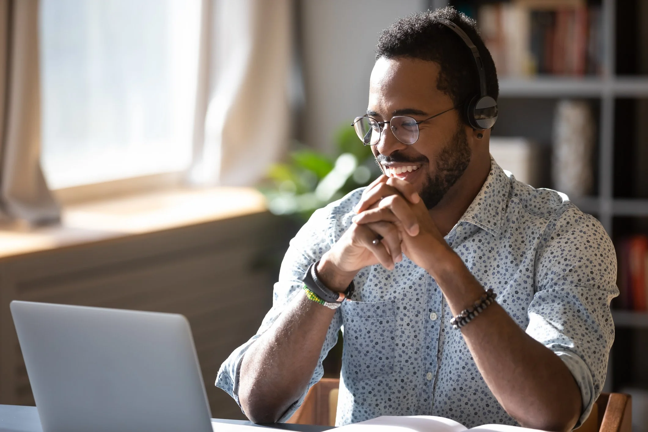 A smiling man with curly hair and glasses, wearing headphones and a patterned shirt, sitting at a desk with a laptop, in a well-lit room with bookshelves and curtains.