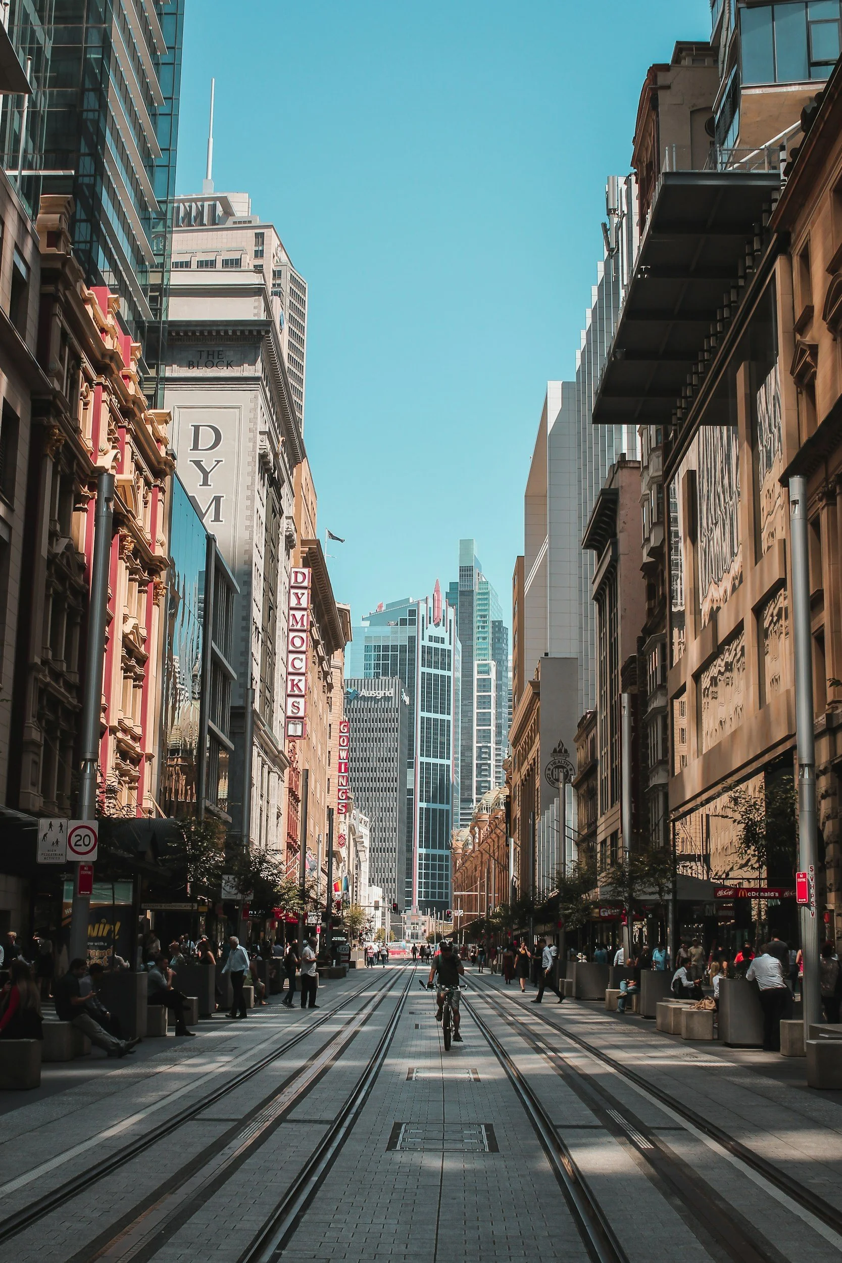 Urban street scene with skyscrapers, a cyclist riding down tram tracks, and pedestrians on the sides, in a city with clear blue skies.