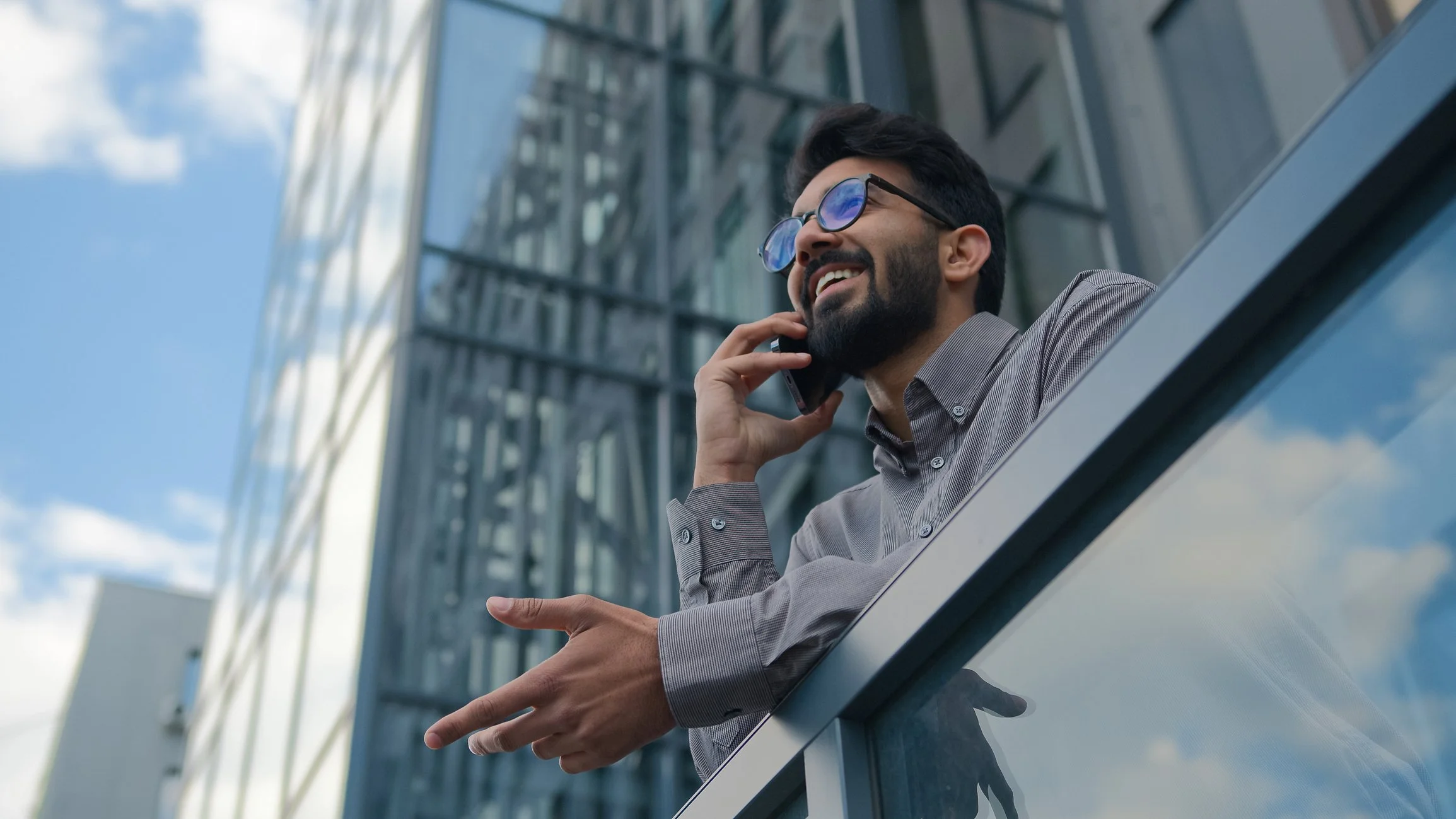 Man with glasses, beard, and gray dress shirt talking on cell phone on balcony with reflective glass building and blue sky in background