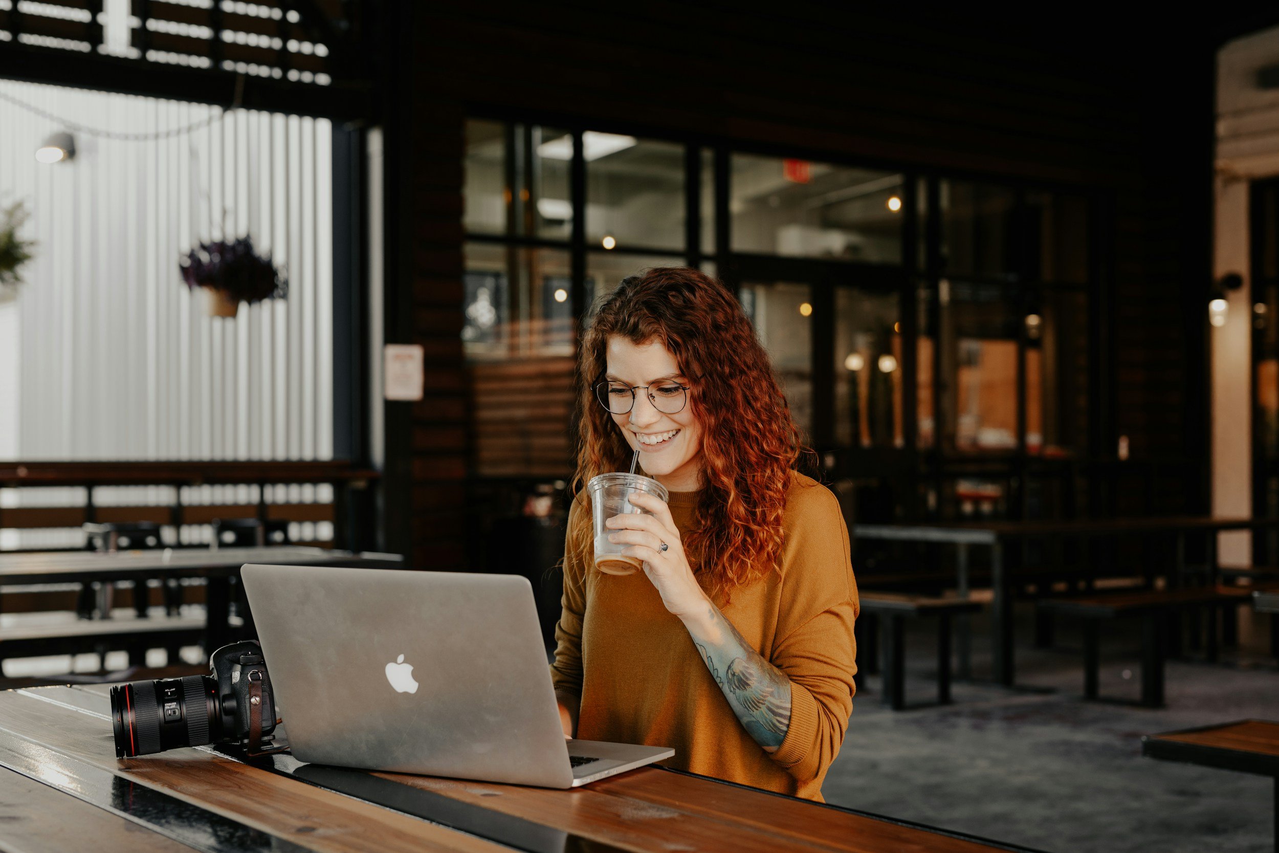 A woman with curly red hair and glasses sitting at a wooden table in a coffee shop, working on a laptop, holding a drink, with a camera on the table beside her.