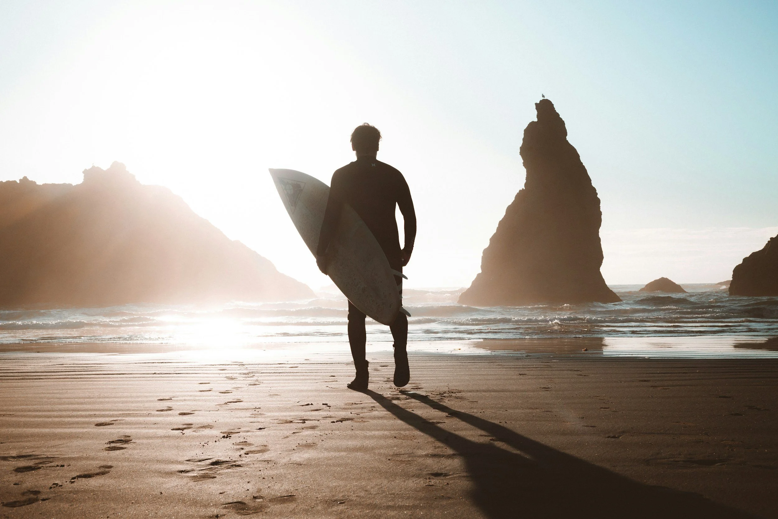 A person with a surfboard walking toward the ocean on a beach during sunset, with large rock formations in the background.