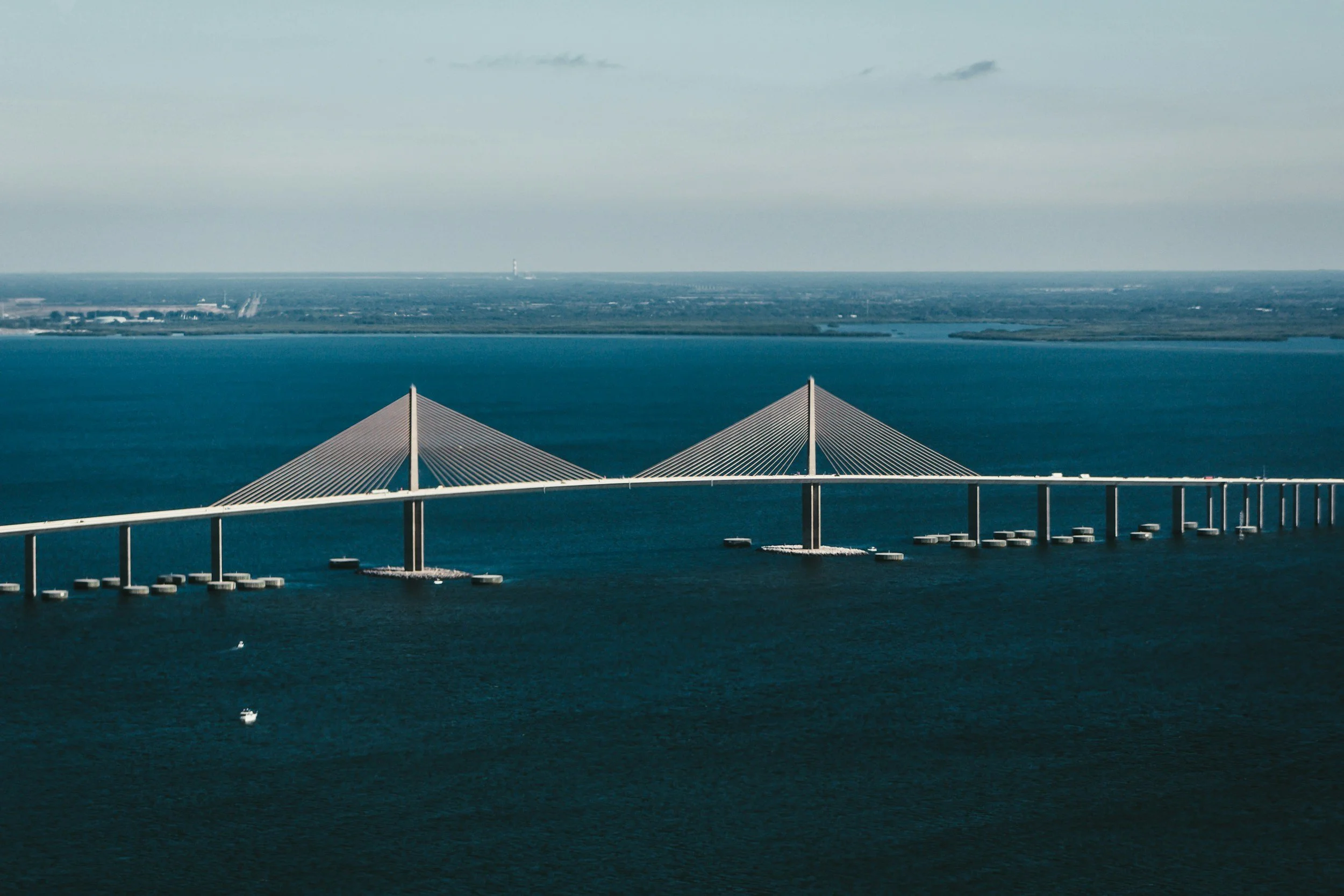 A large bridge with cable-stayed design extending over a body of water, with two prominent pyramid-shaped pylons supporting the cables, and a distant shoreline in the background.