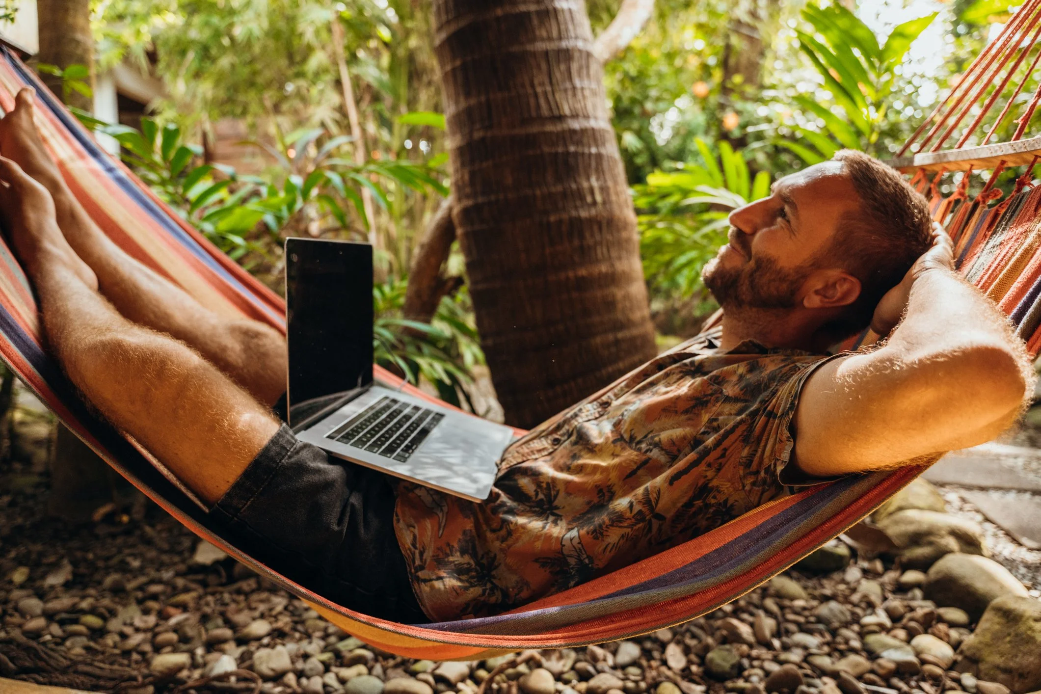 A man relaxing in a hammock outdoors with a laptop on his stomach, surrounded by tropical plants and trees.