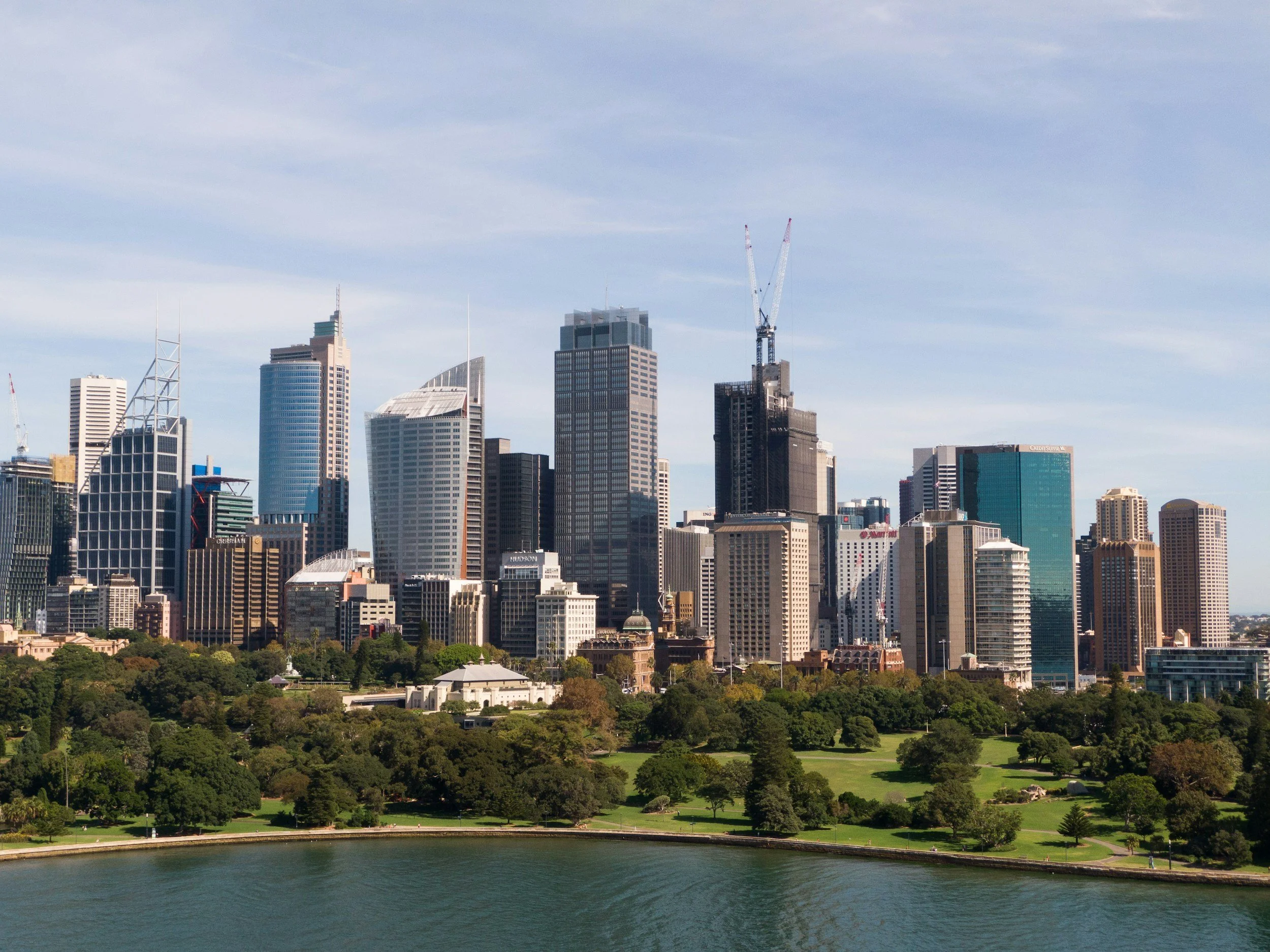 A city skyline with tall modern skyscrapers and buildings, a construction crane, greenery, and a body of water in the foreground.