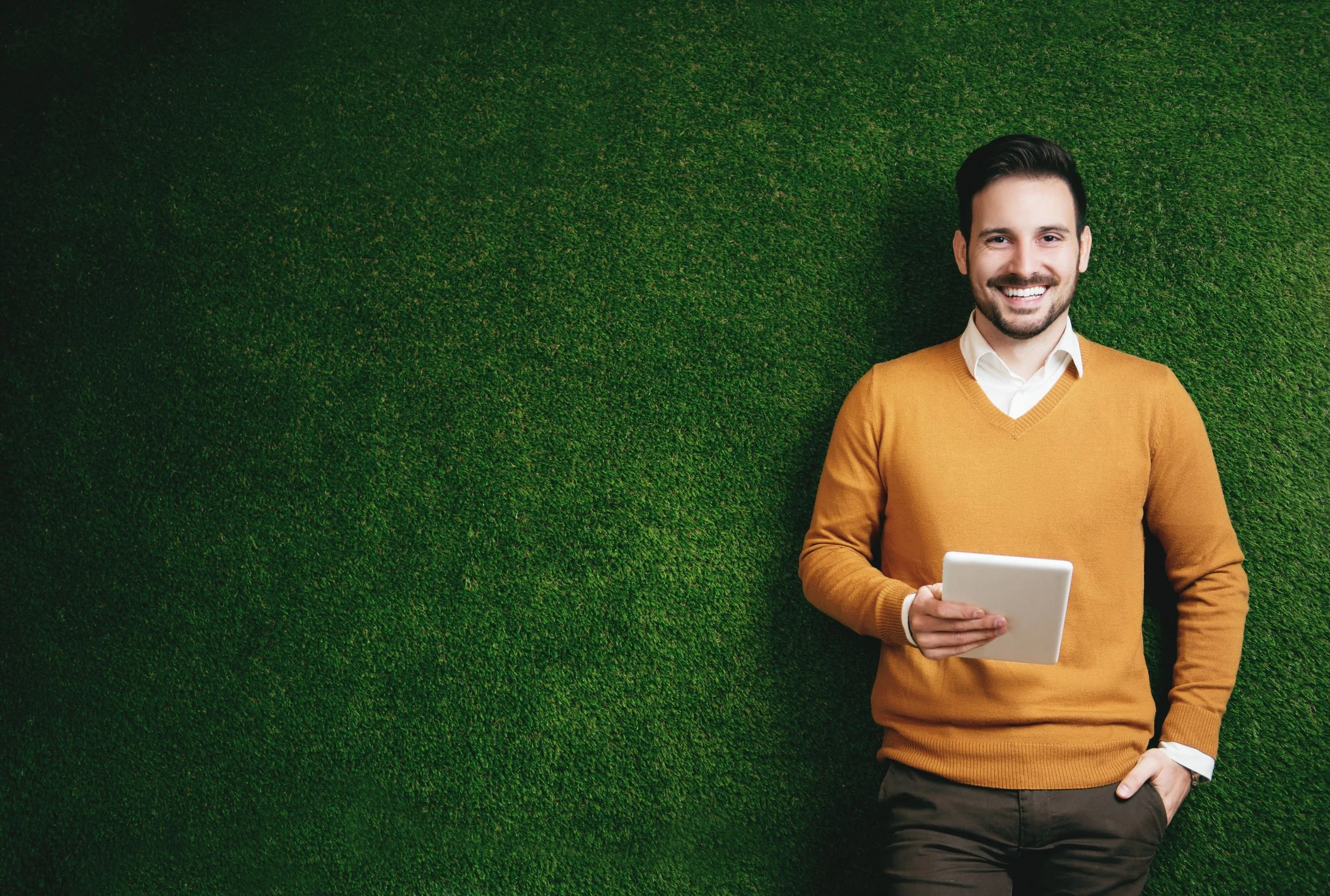 A young man with dark hair, a beard, wearing a mustard-colored sweater over a white shirt, holding a tablet, standing against a green textured wall, smiling.