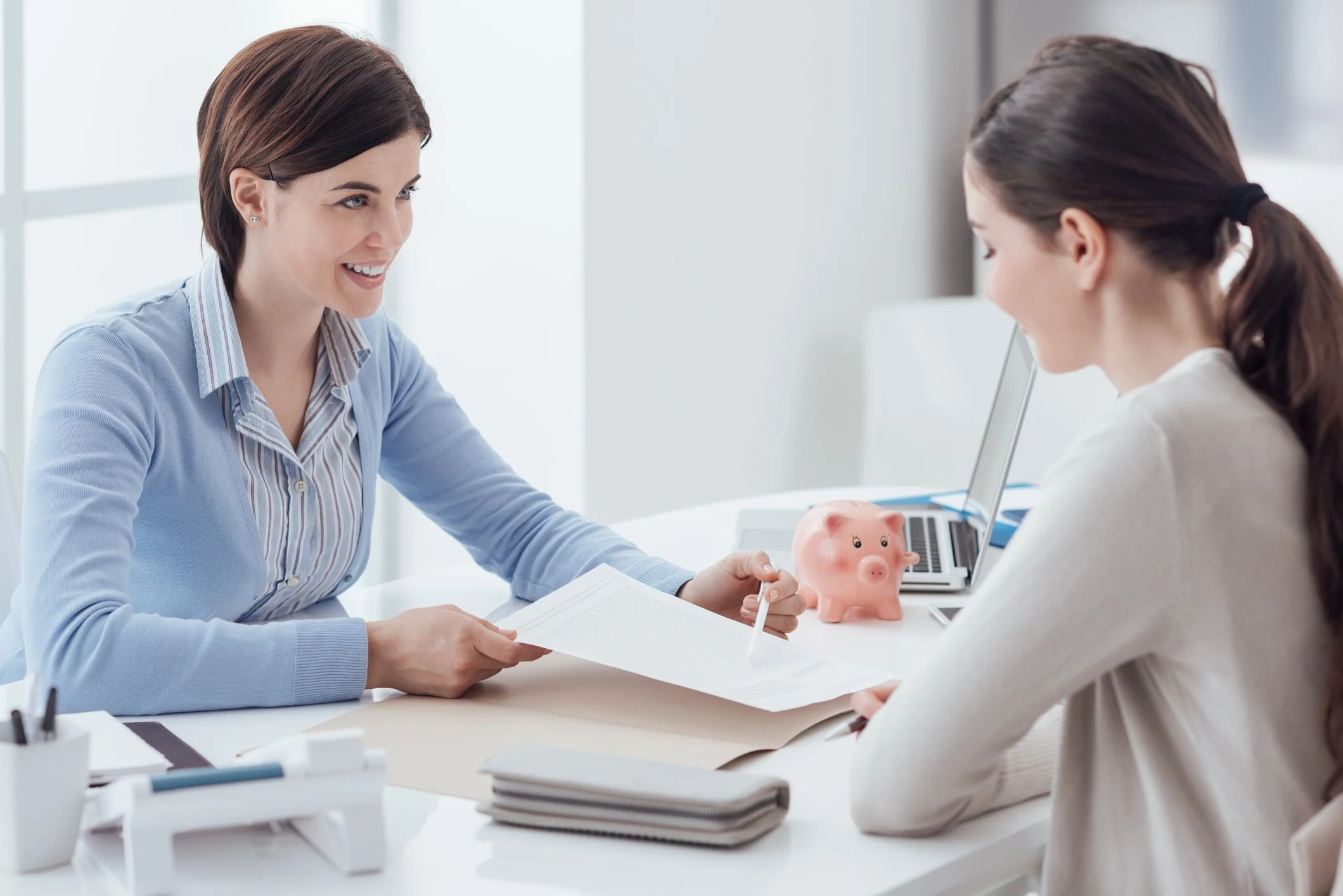 A woman at a desk talking to another woman. The woman on the left has short brown hair and is smiling, holding a document. The woman on the right has long brown hair tied back, wearing a white sweater. There is a pink piggy bank, a laptop, and office supplies on the desk.