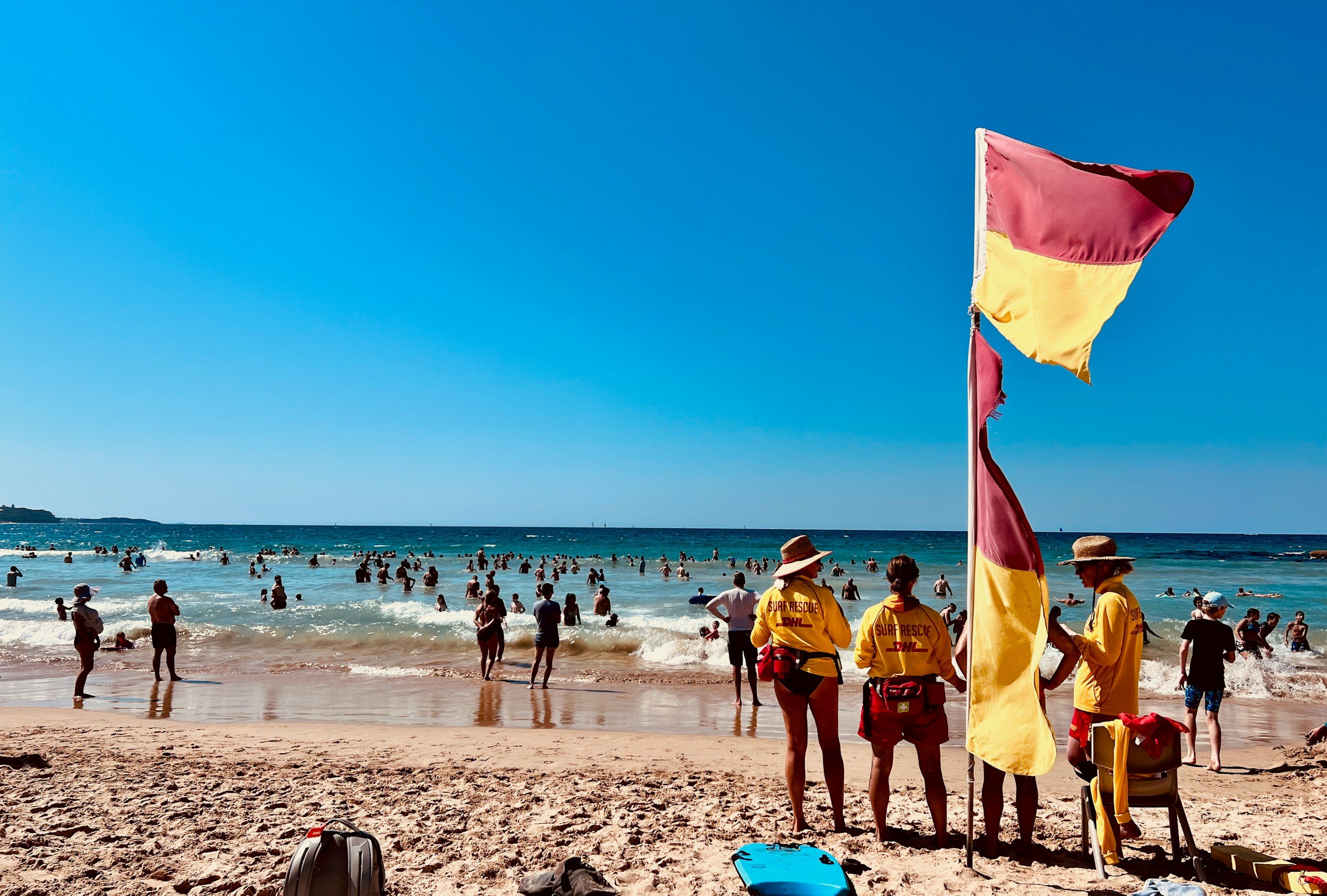 Beach scene with many people swimming and relaxing in the ocean. In the foreground, four surf rescue personnel in yellow uniforms and hats stand near a flag on a pole, with rescue equipment on the sand.