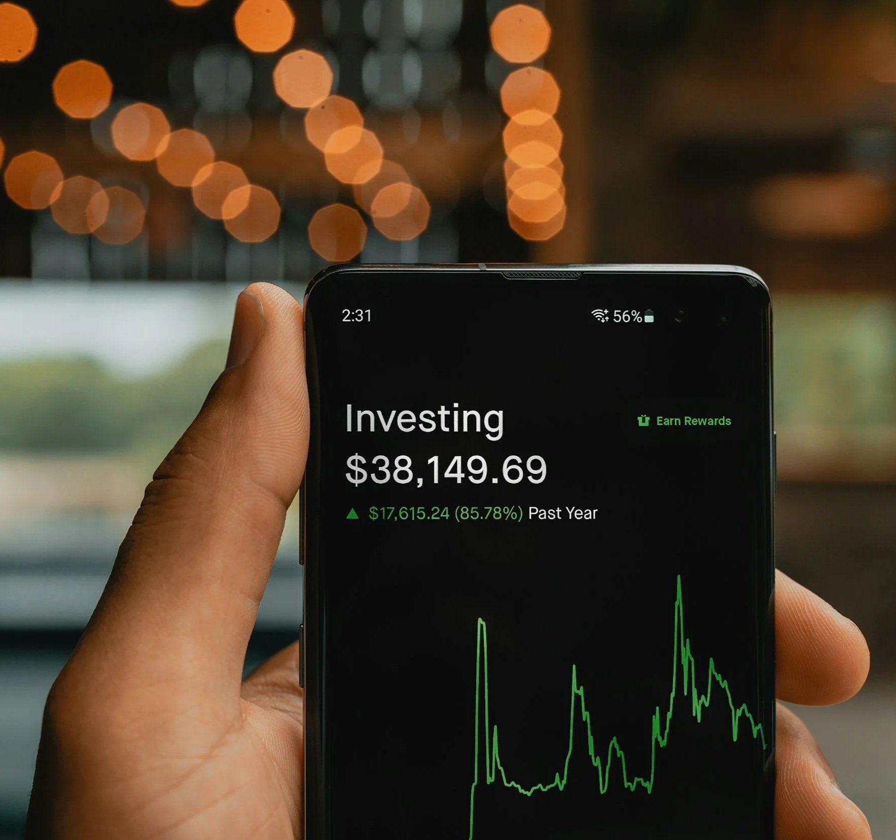 A person holding a smartphone displaying a financial investment app with a portfolio value of $38,149.69, up by 85.78% over the past year, with a line graph below showing investment fluctuations.
