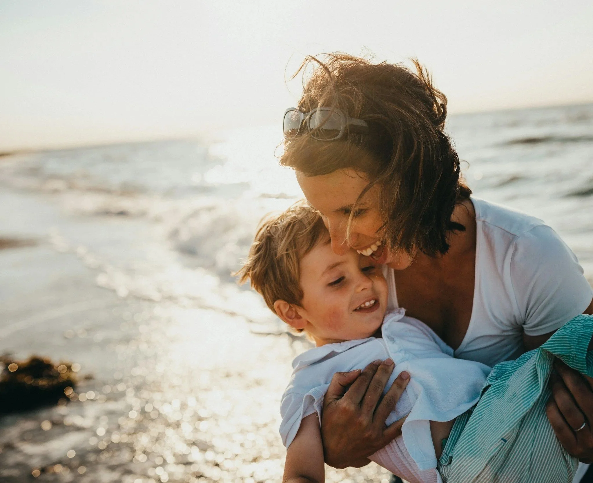 A woman and a young boy playing and hugging at the beach during sunset.