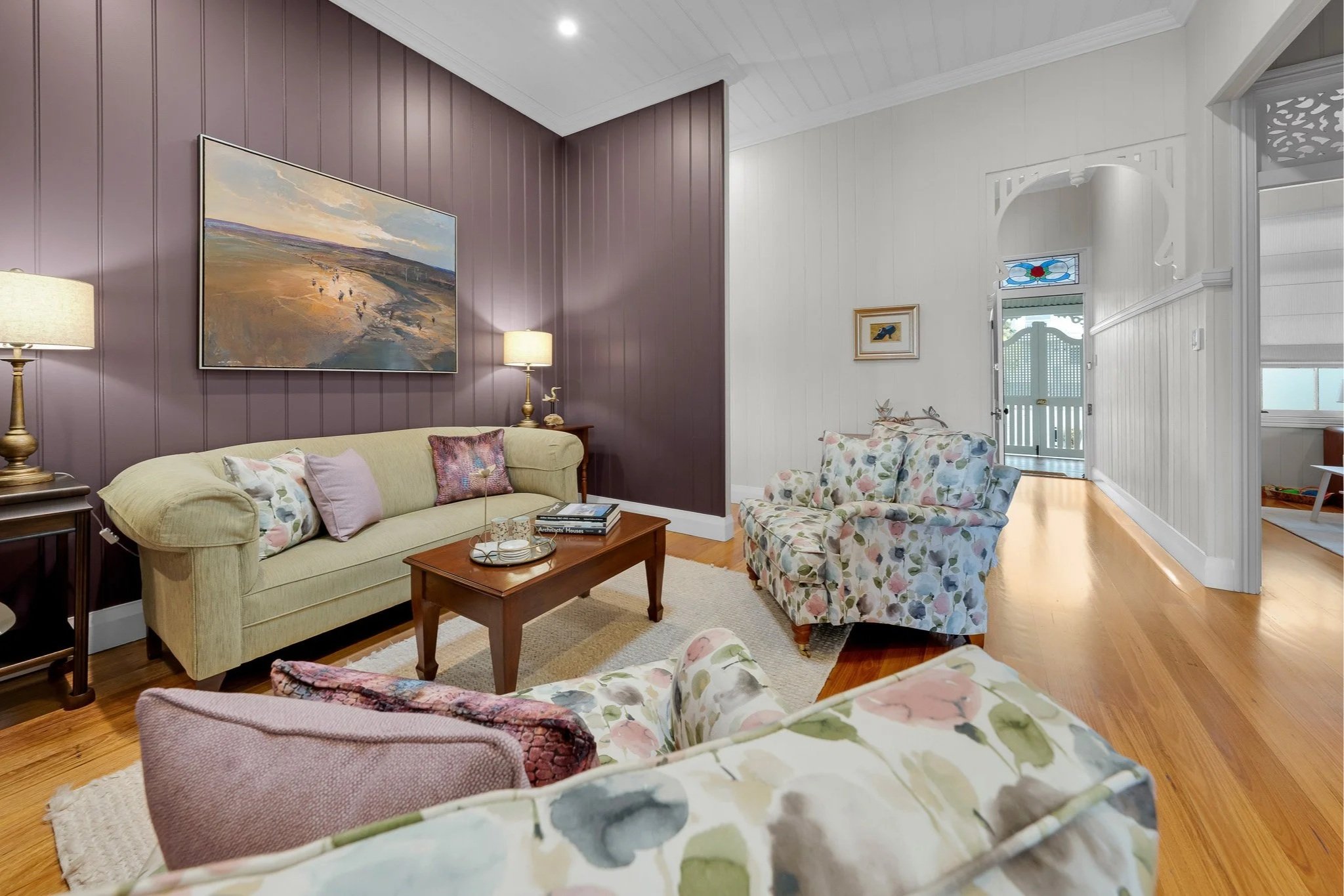 A cozy living room with floral-patterned armchairs, a green sofa with decorative pillows, a wooden coffee table, and purple and white paneled walls. There's a landscape painting above the sofa and lamps on side tables. A door with a stained glass win
