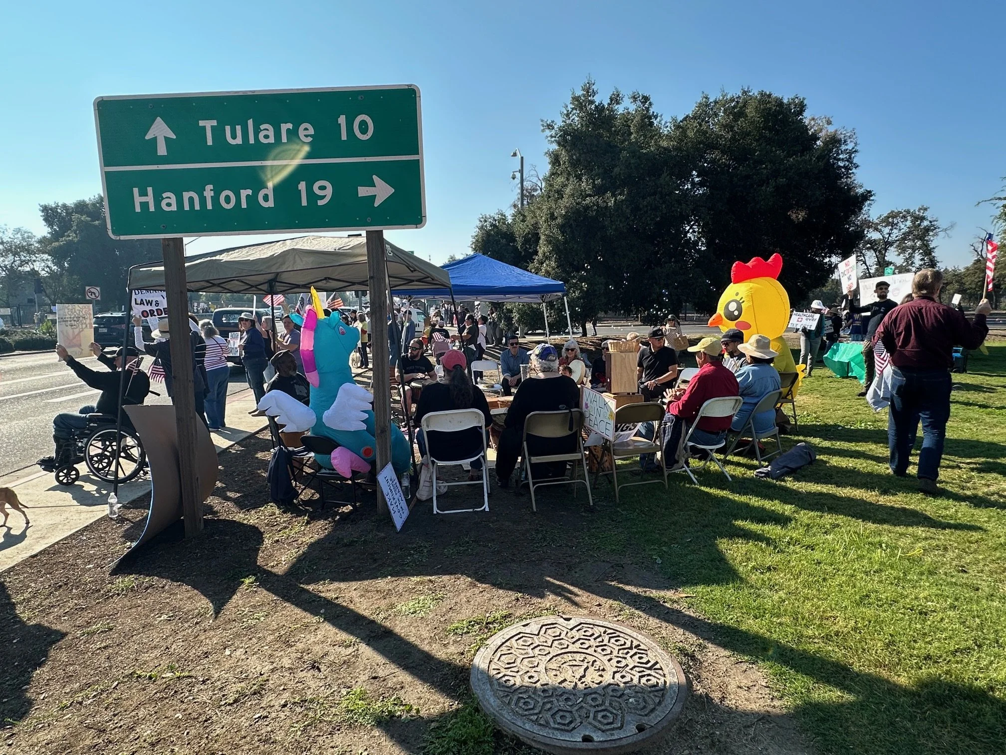 A group of people gathered outdoors at a rally or protest with some dressed in costumes, including a unicorn and a rubber duck, under tents and near signs, with a green highway sign indicating directions to Tulare and Hanford, California.