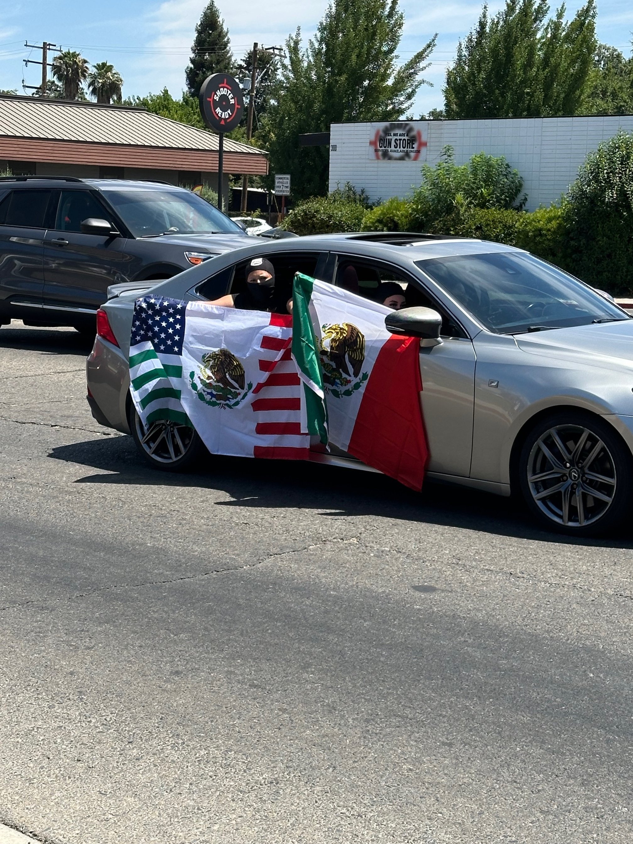 A silver car with the Mexican flag and the American flag draped over its side, parked on a street with a person visible inside wearing a black face mask and hat, with another person visible through the window. There are trees, a sign for a gun store,