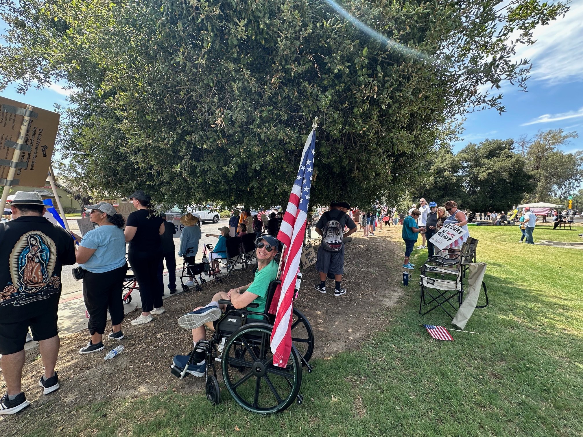 A woman sitting in a wheelchair holding an American flag at a gathering under a large tree, with many people standing and sitting nearby holding signs and flags.