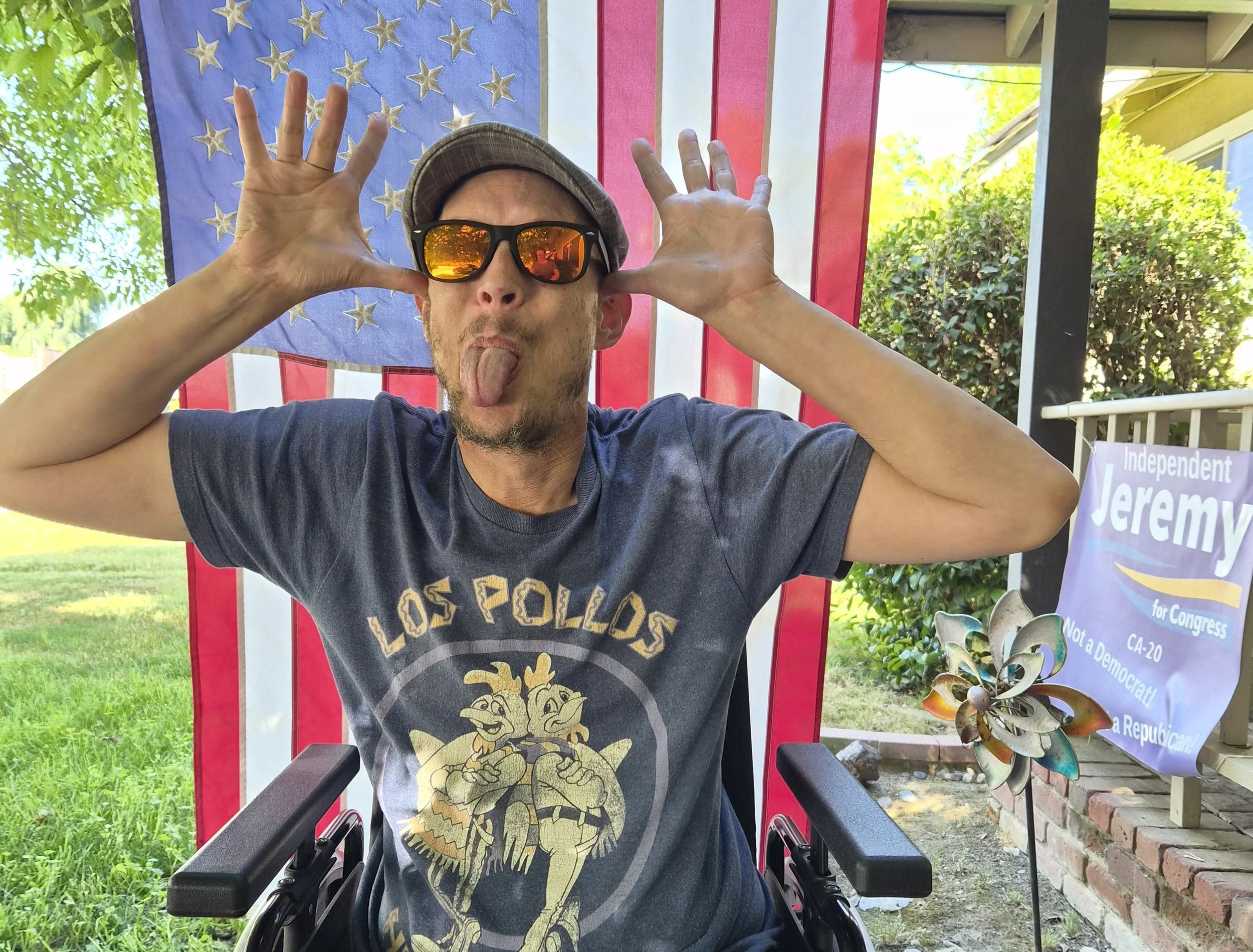 A man wearing sunglasses and a baseball cap blowing a kiss while sitting in a wheelchair in front of an American flag backdrop.