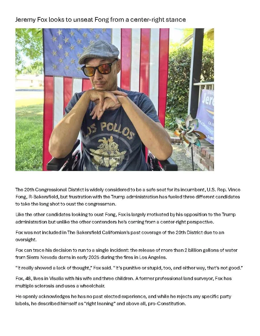 Jeremy Fox wearing sunglasses and a cap sitting in front of an American flag, with a pinwheel on a table nearby, appearing to look downward with his hands clasped. Bakersfield Californian