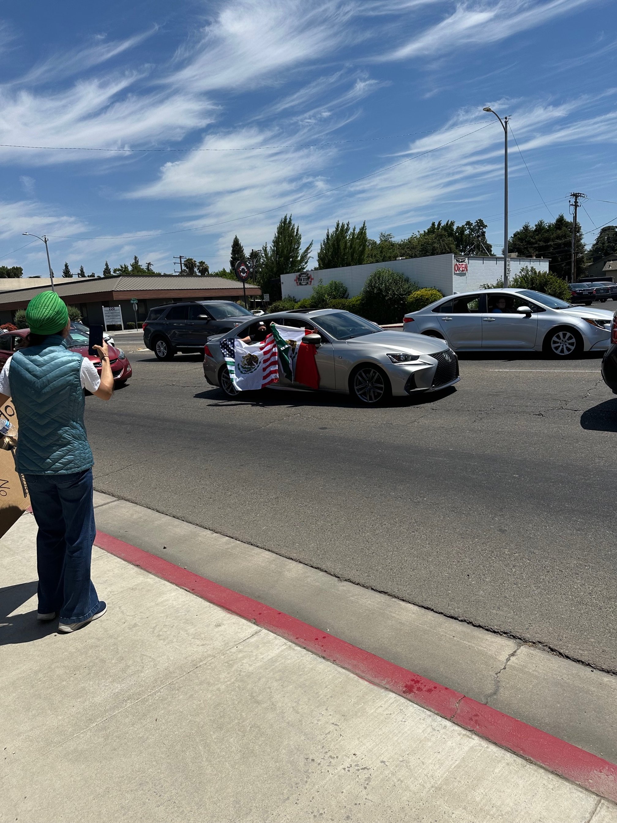 A silver car decorated with Mexican flags and a red scarf, participating in a parade or celebration, on a sunny day with a woman taking a photo on the sidewalk.