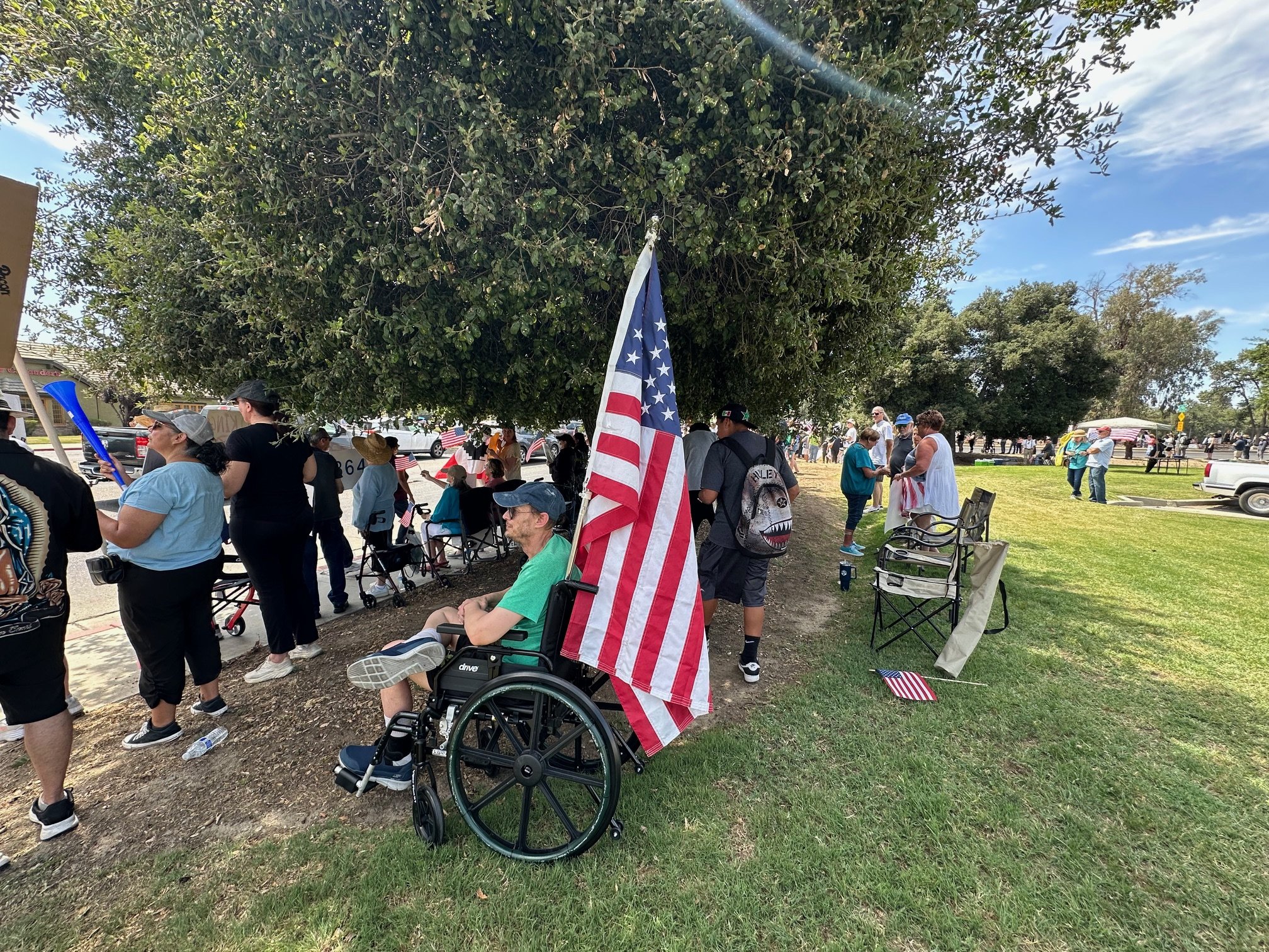 A gathering of people in a park with many American flags, some sitting and some standing, under large trees on a sunny day.
