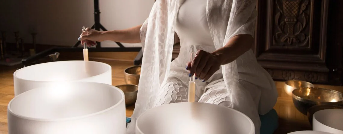 Person playing crystal singing bowls, wearing white, kneeling on a cushion on the floor.