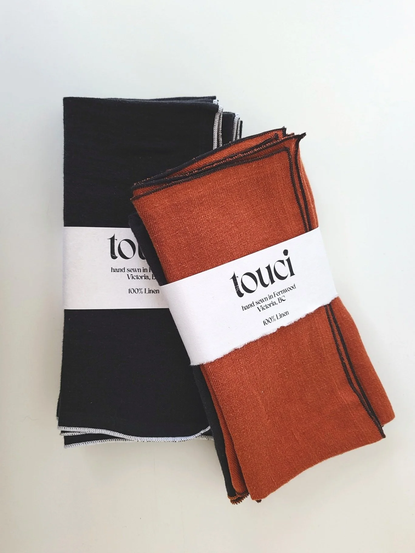 Two folded linen cloths, one black and one orange, with branding labels wrapped around each indicating they are handmade from 100% linen.