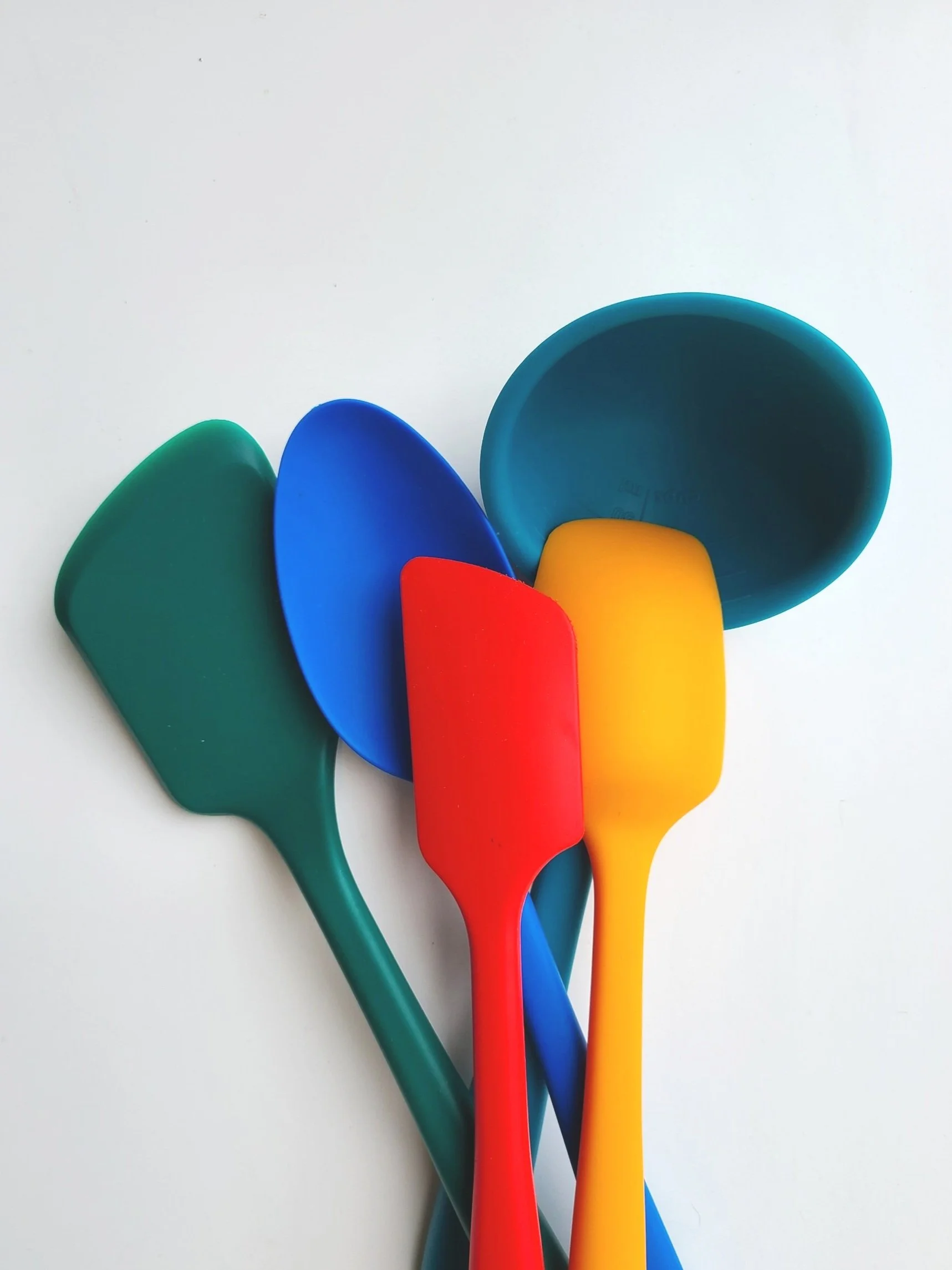 Colorful plastic kitchen utensils, including a green spatula, blue spoon, red spatula, yellow spoon, and teal bowl, arranged on a white surface.