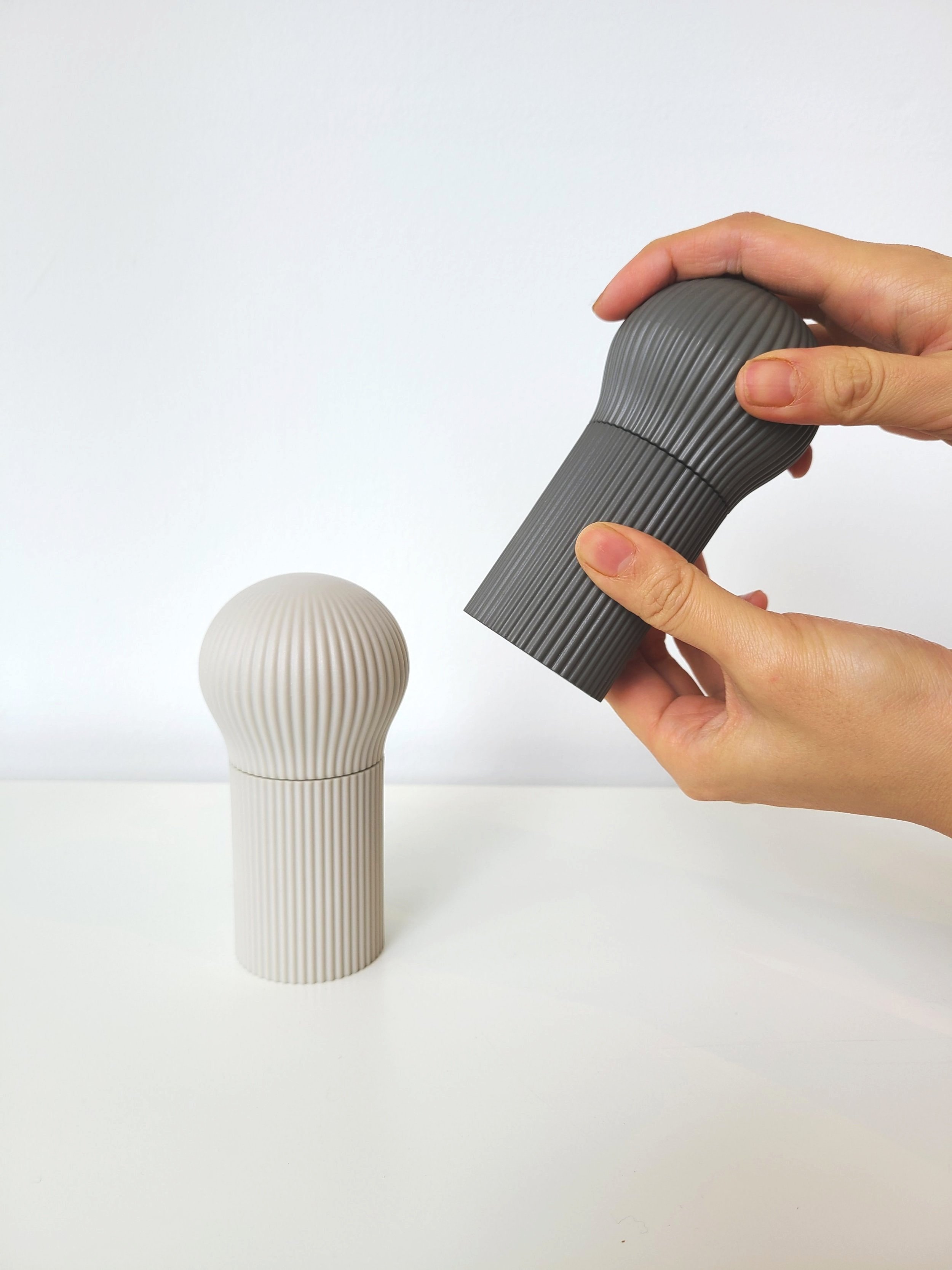 Person holding a dark gray, ribbed salt or pepper grinder above a matching light gray grinder on a white surface with a plain white background.