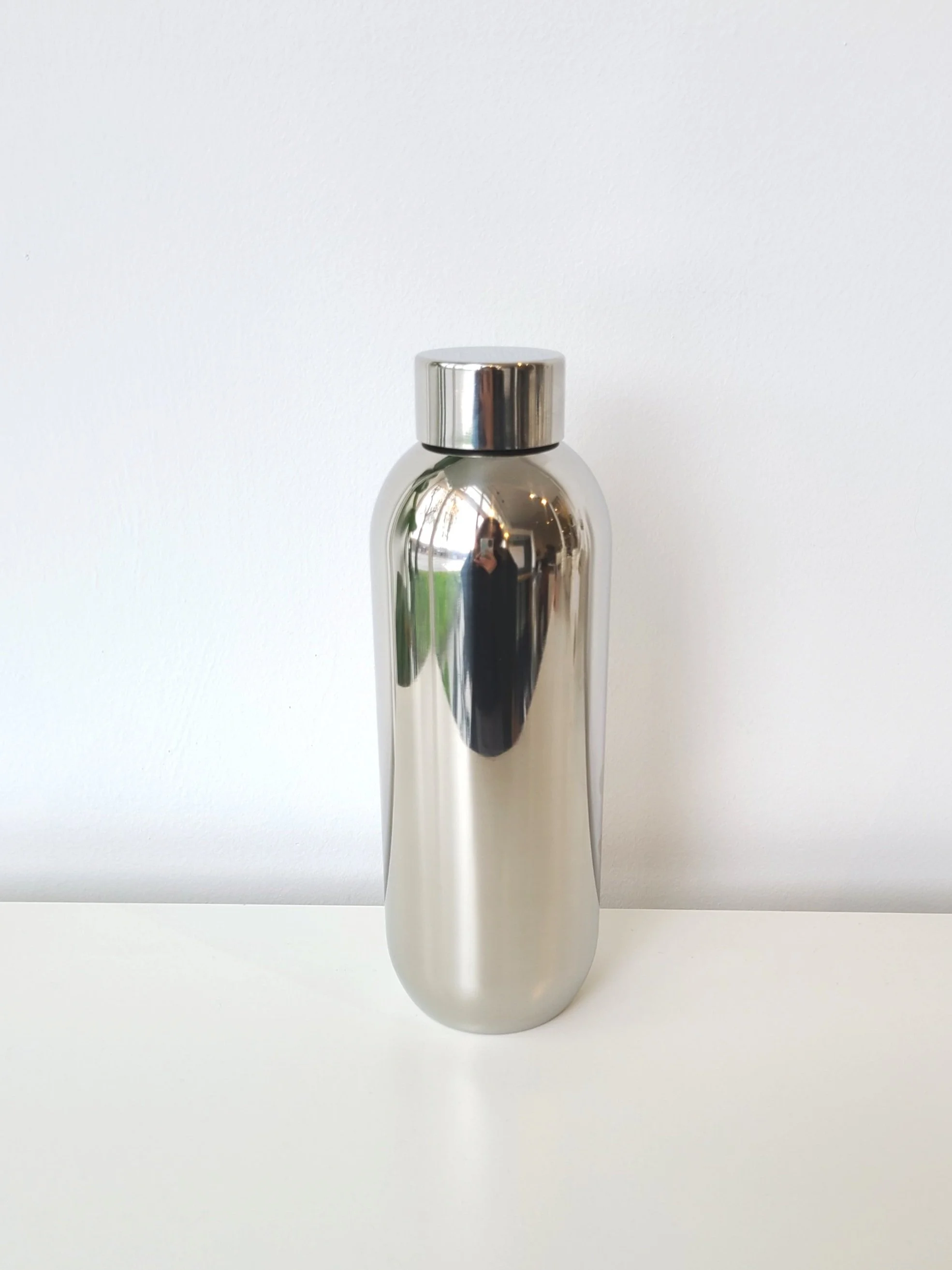 A metallic, reflective, stainless steel water bottle with a silver cap, placed on a white surface against a white background.