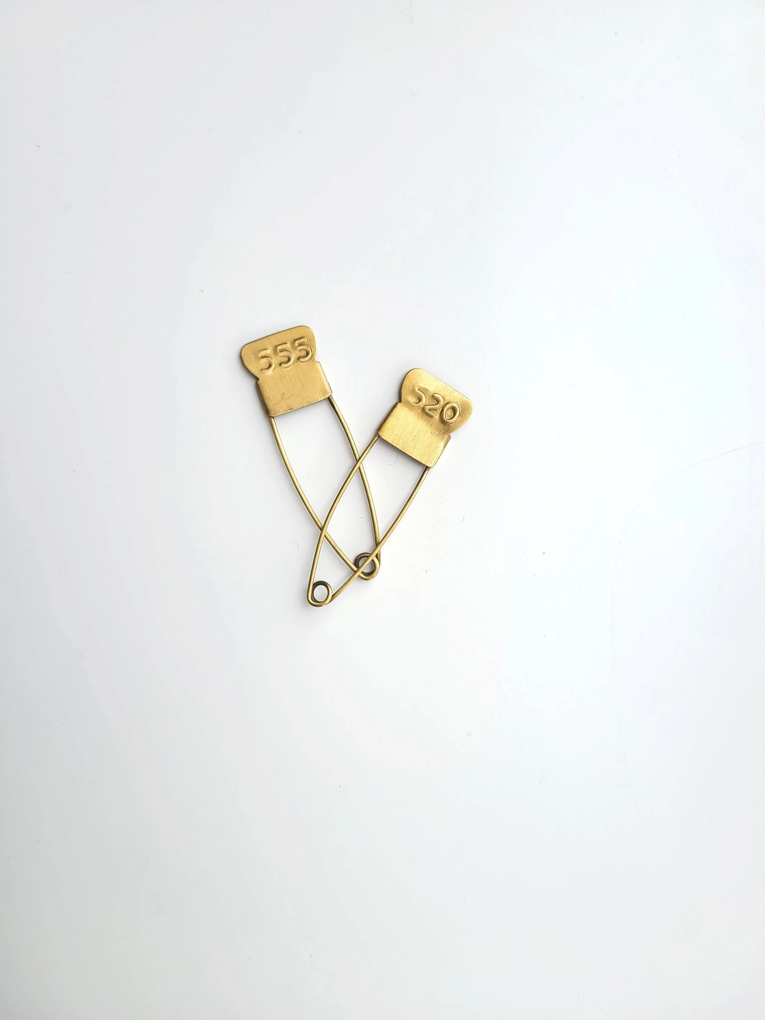Two gold-colored safety pins with engraved numbers '555' and '520' lie crossed on a white background.