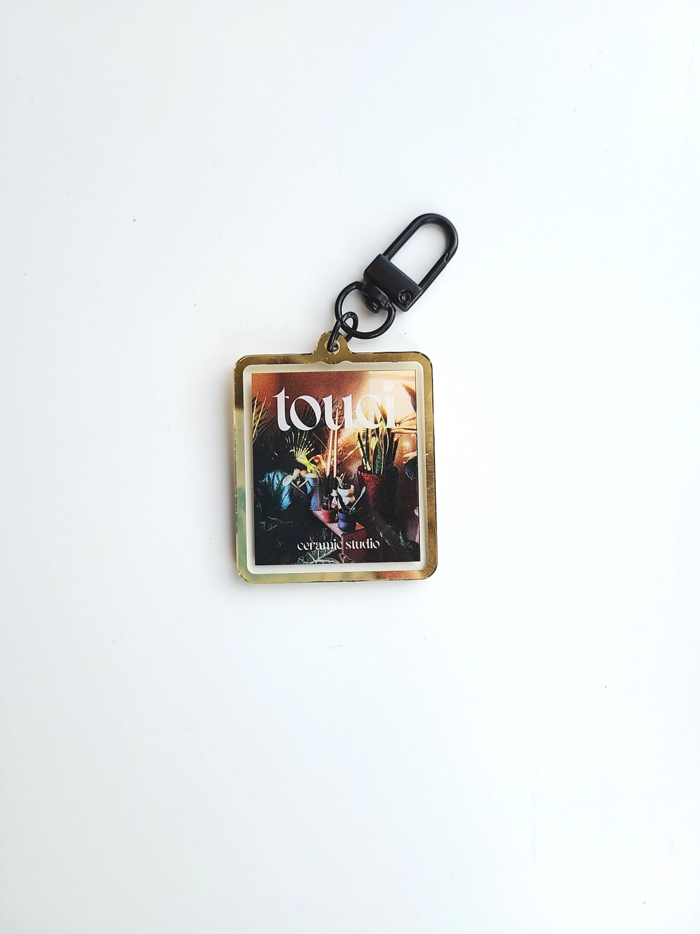 A small rectangular keychain with a black clip, featuring an image of colorful potted plants and the words 'touch ceramic studio' on a white background.