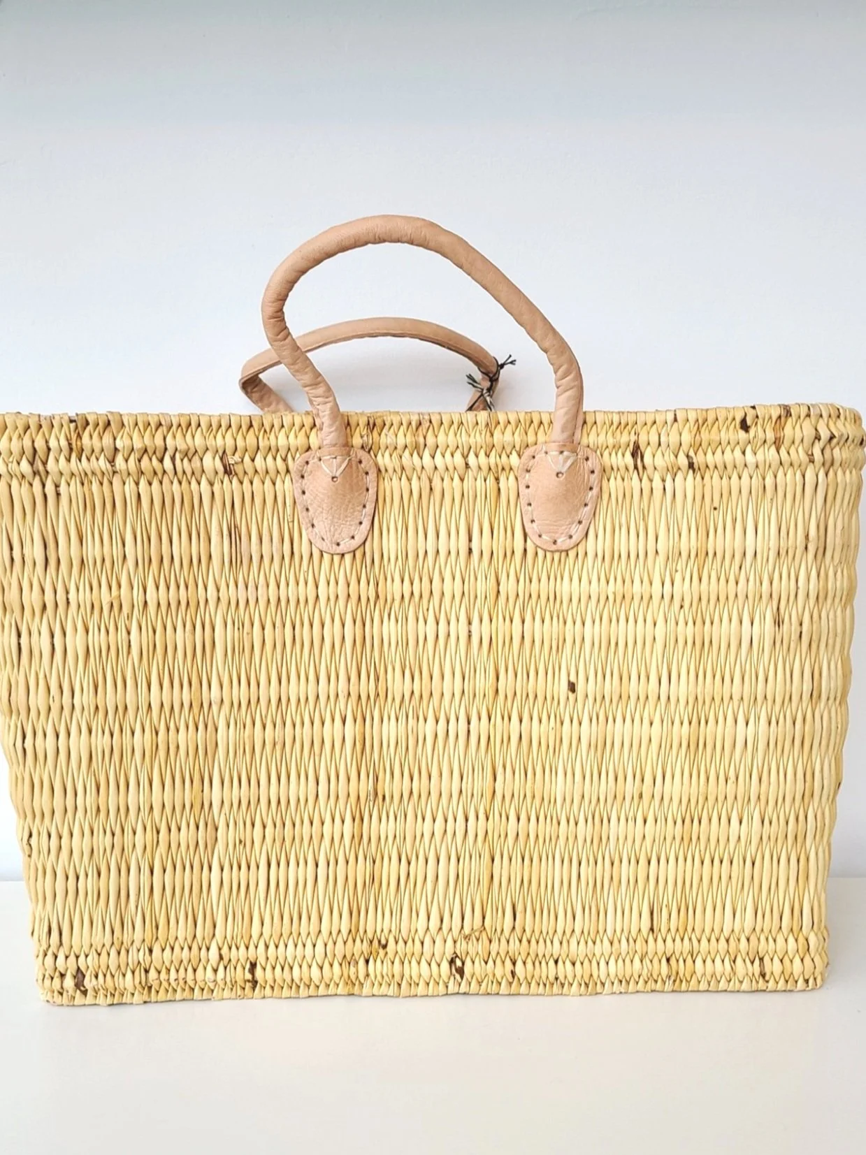 Tan woven straw tote bag with leather handles and accents against a light gray background.