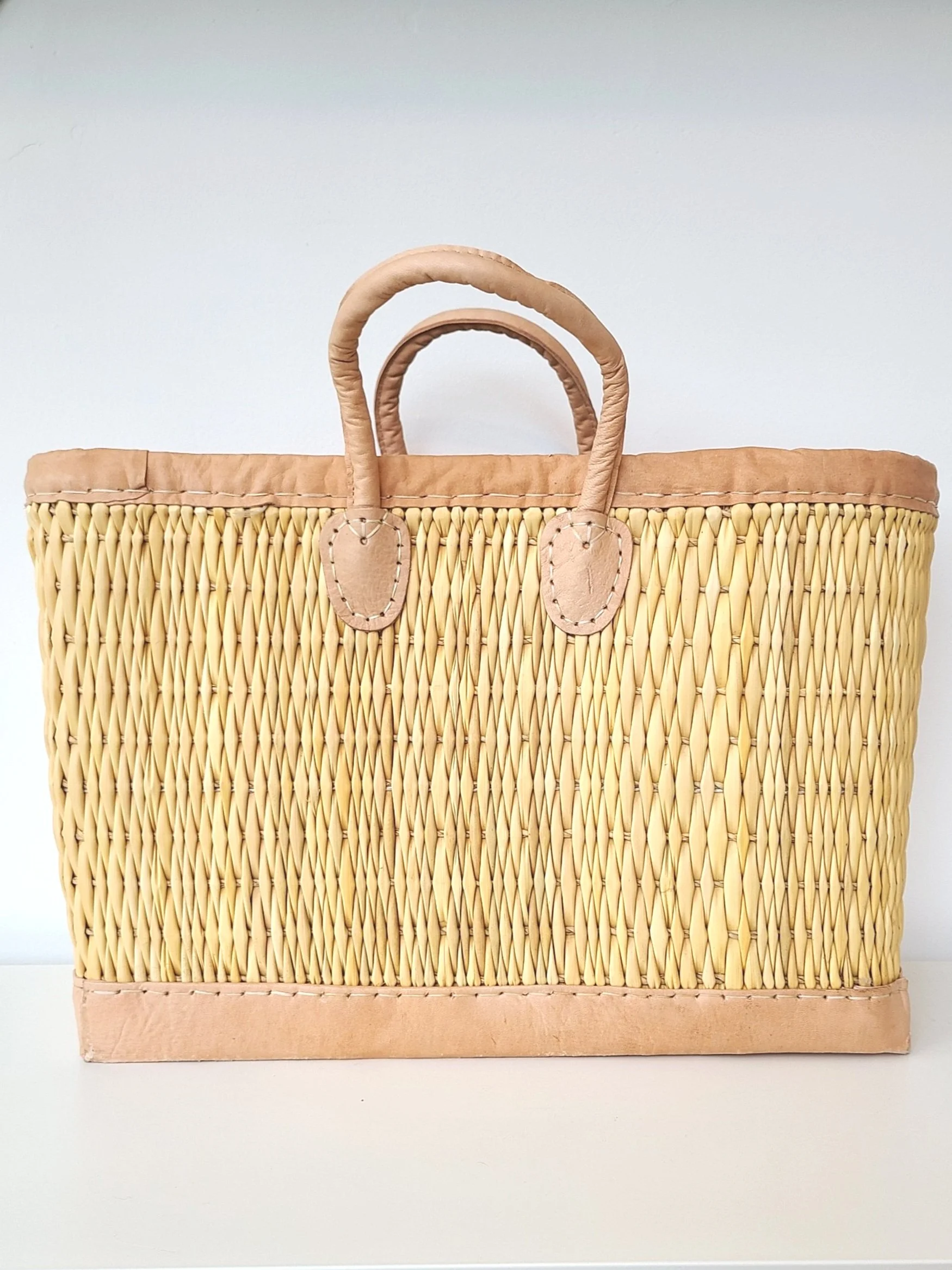A woven straw handbag with leather handles and trim, placed on a white surface against a plain background.