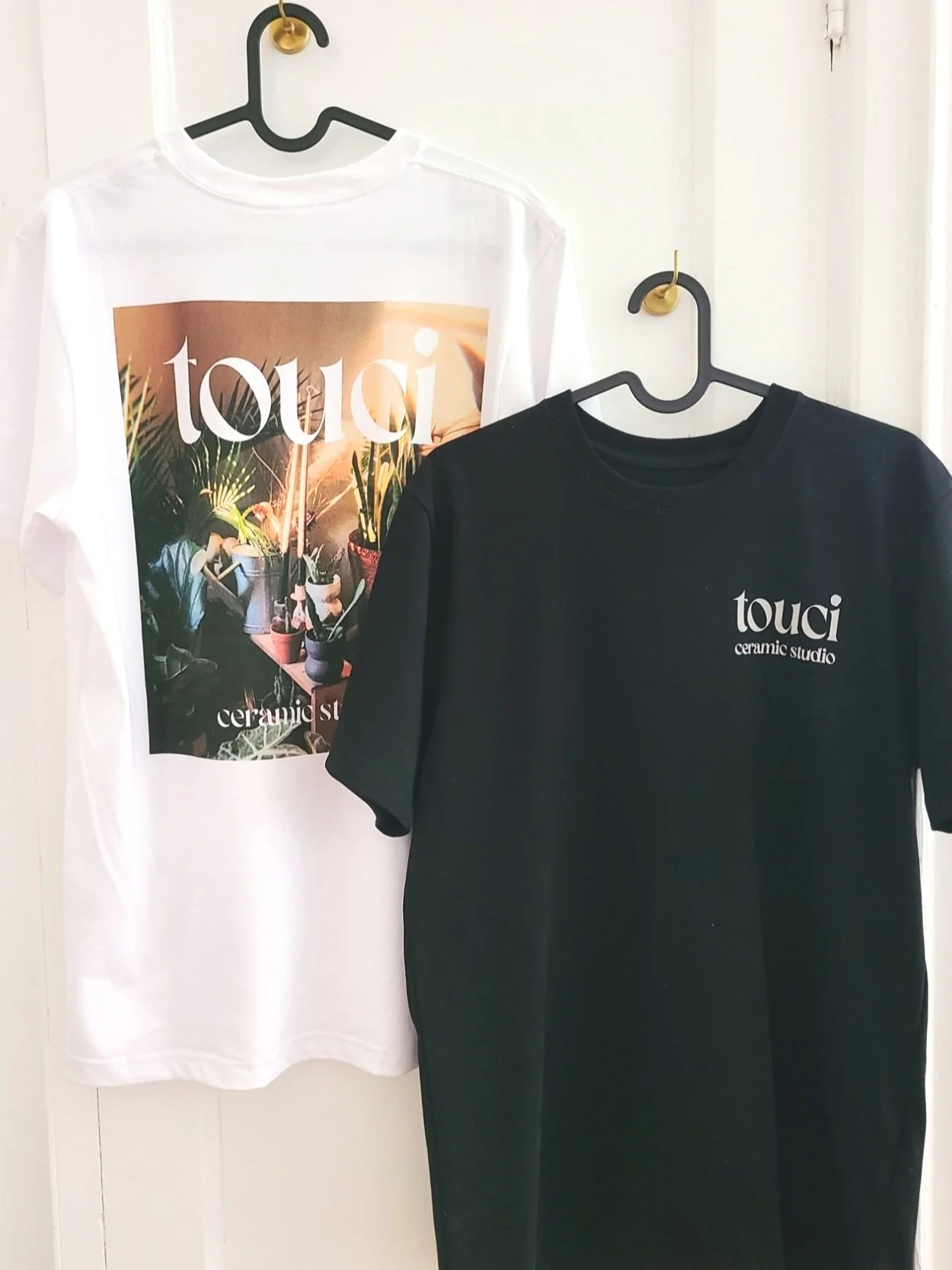 Two shirts hanging on hooks; one white with a graphic of potted plants and the word 'toui,' and one black with the text 'toui ceramic studio'.