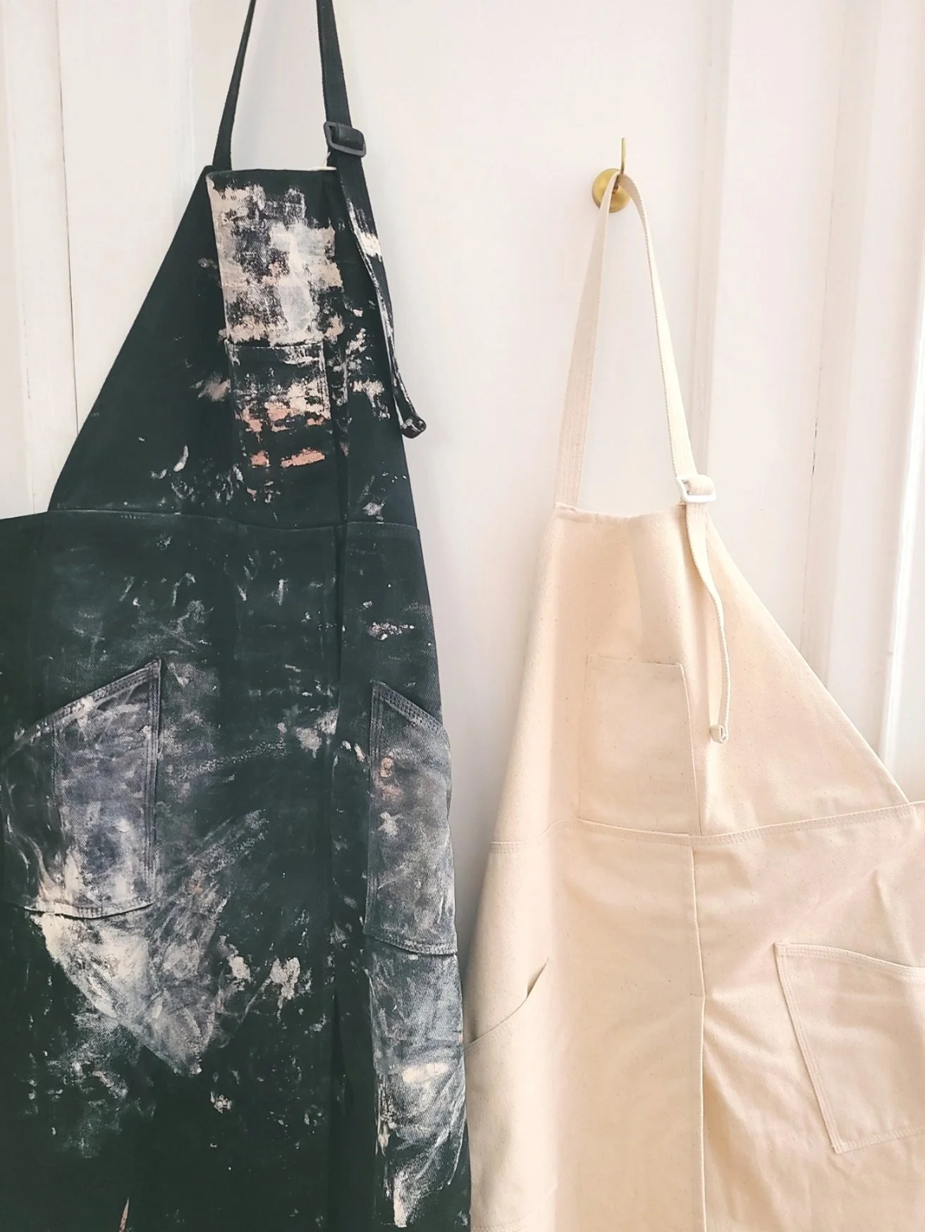 Two aprons hanging on hooks against a white wall: one black with paint splatters and smudges, the other plain beige.
