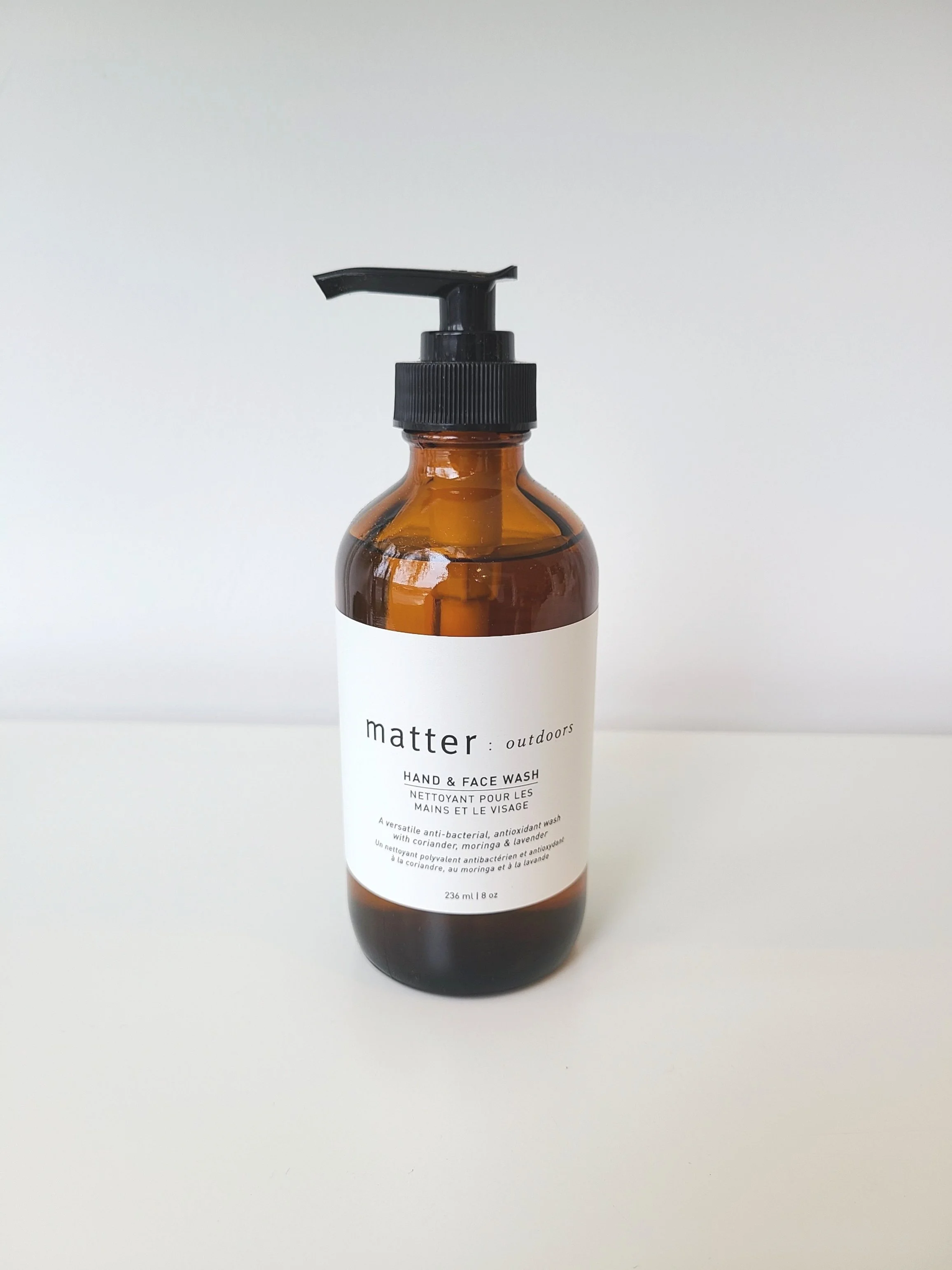 Amber glass bottle of Matter Outdoors hand and face wash with black pump dispenser, placed on a white surface against a plain white background.