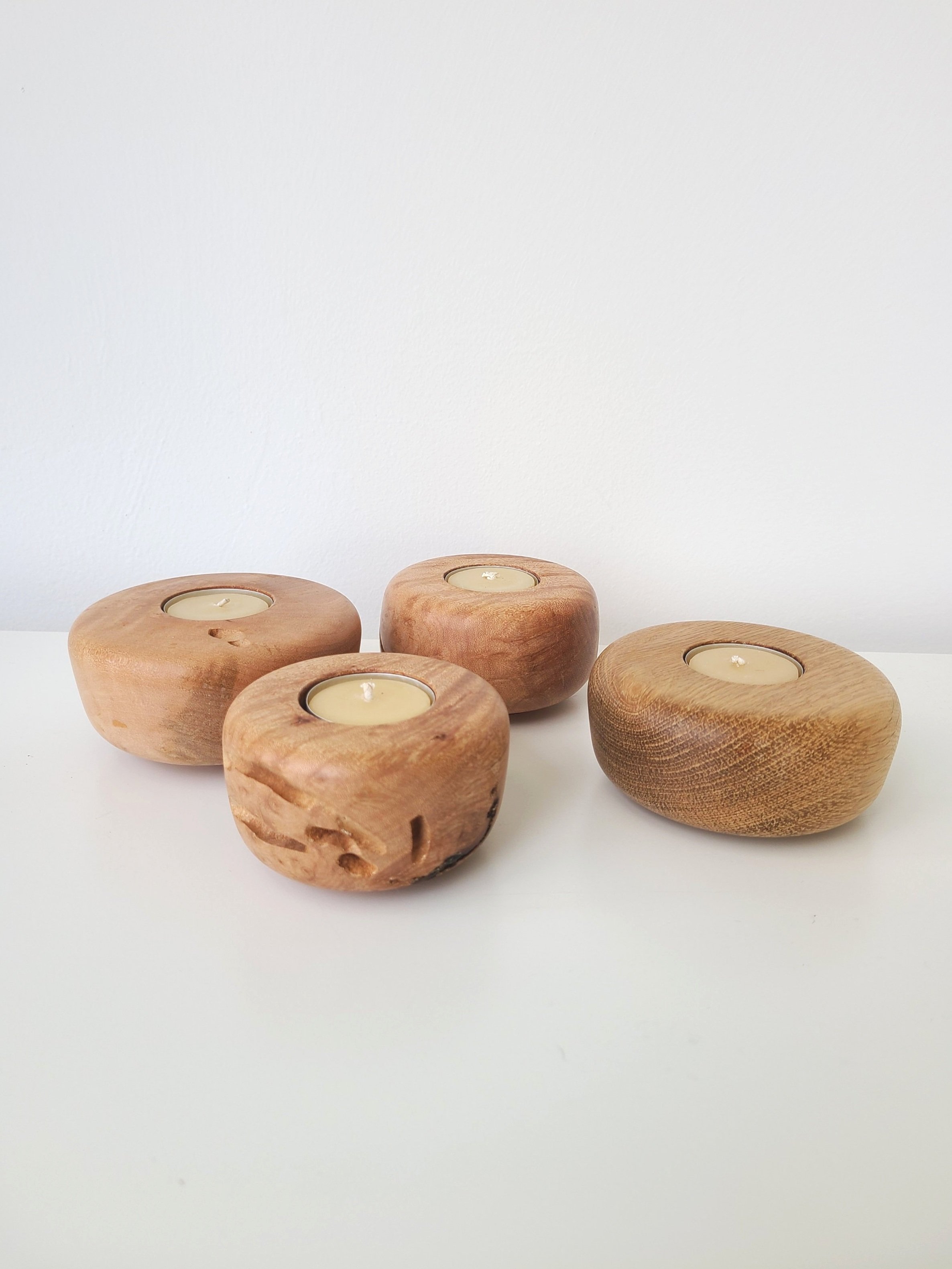 Four wooden candle holders with tea light candles on a white surface against a white wall.