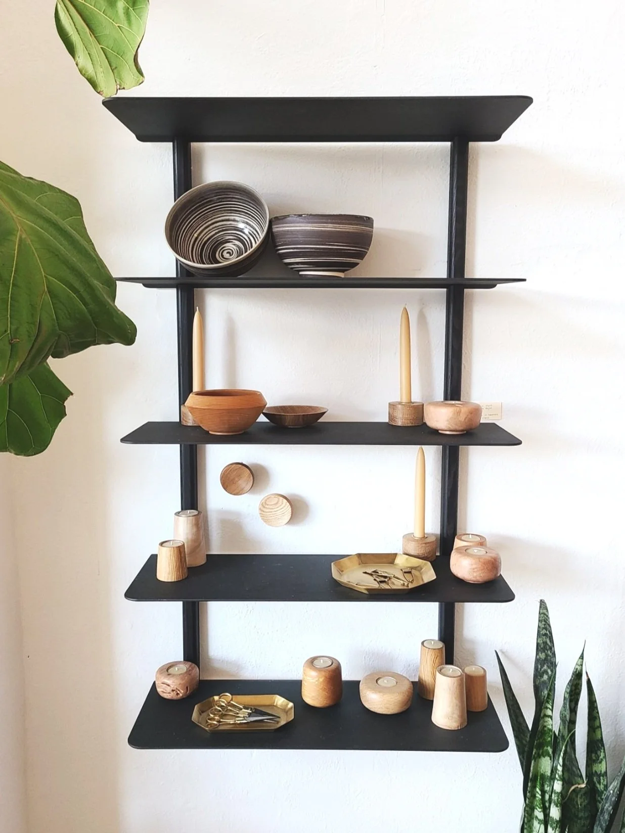 Wall-mounted black shelf unit displaying various wooden and ceramic decorative items, including bowls, candle holders, and a gold tray, with greenery on the left and right sides.