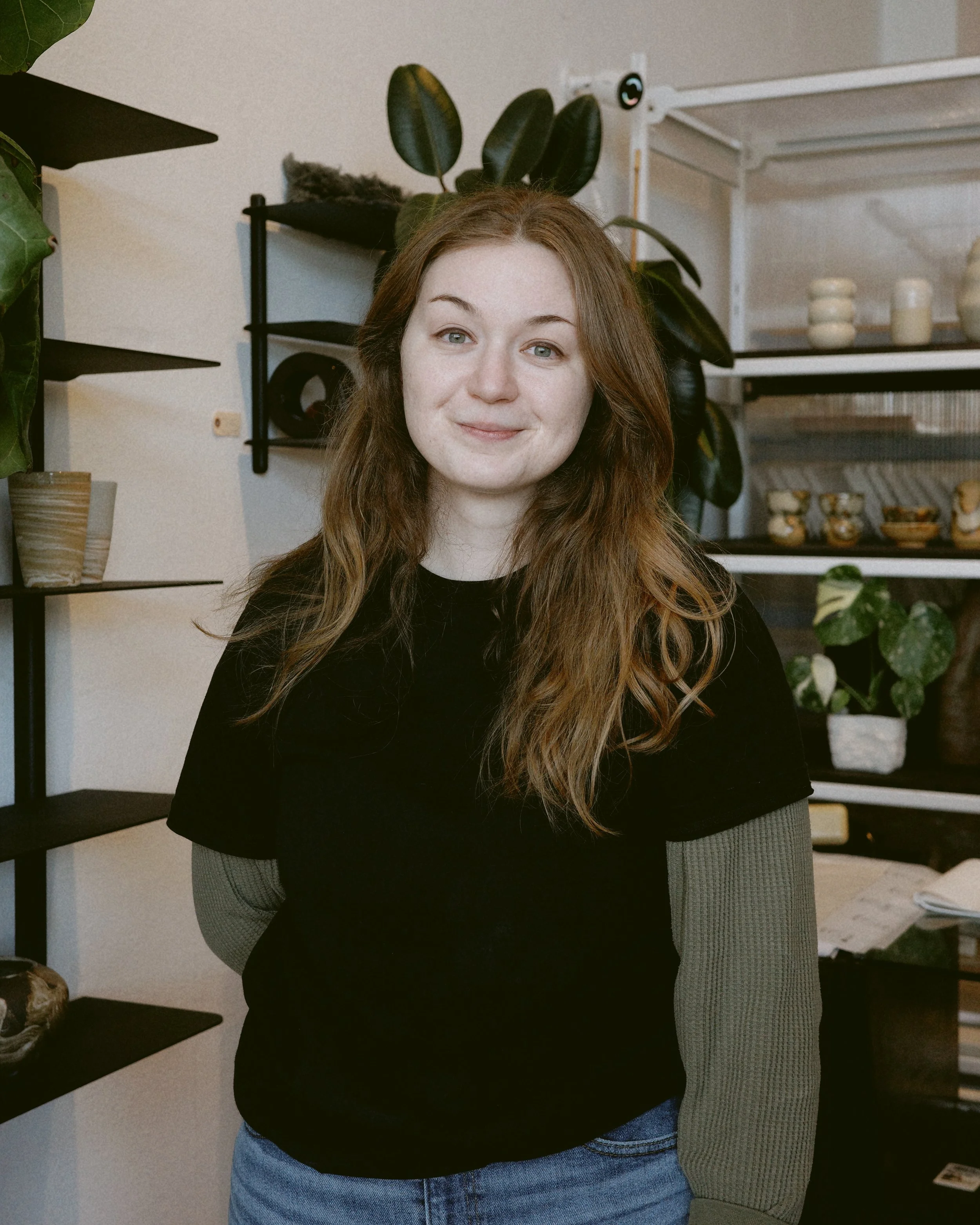 A young woman with red hair standing indoors in front of shelves with plants and dishes, smiling at the camera.