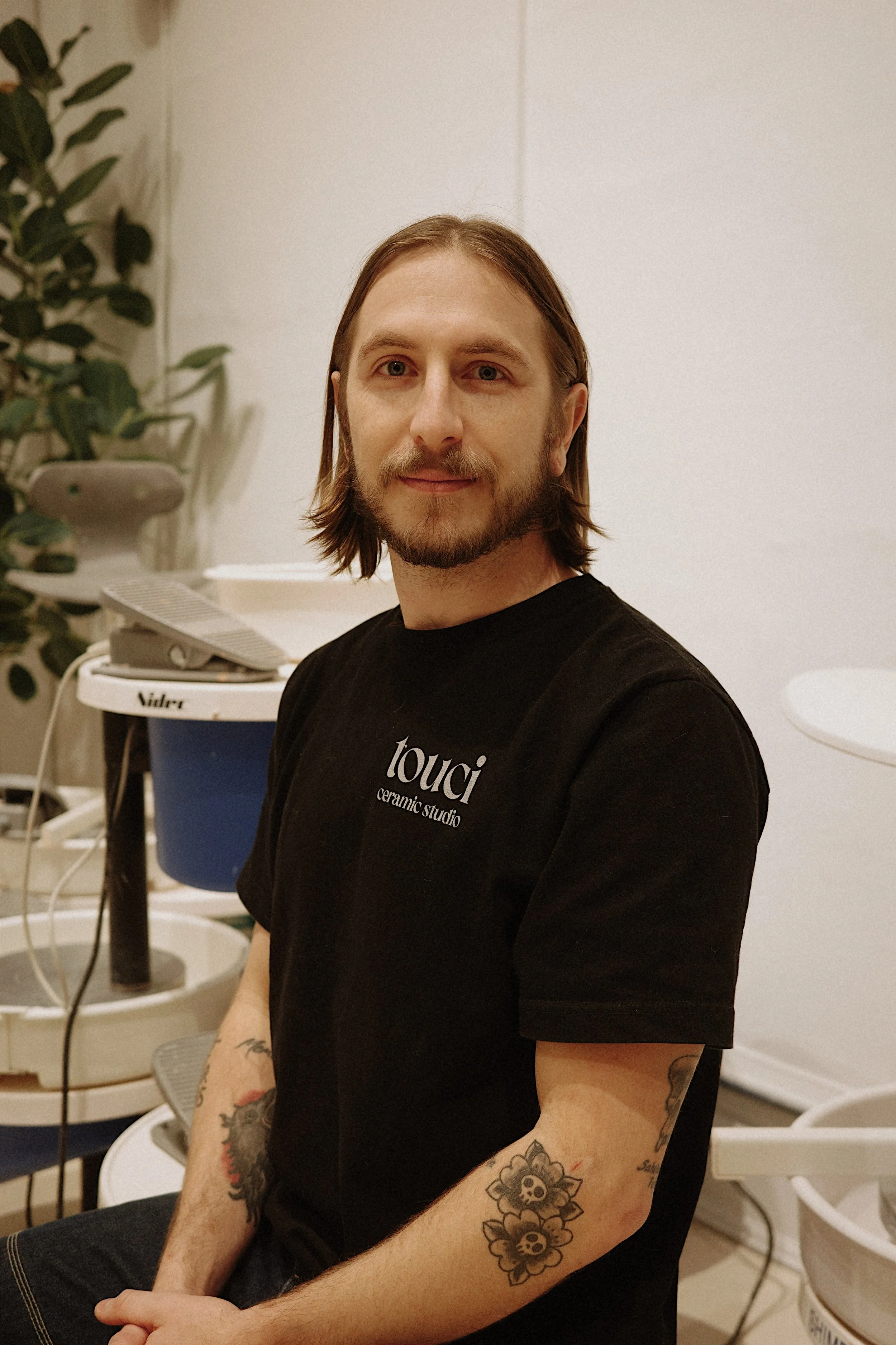 A man with shoulder-length brown hair and a beard, wearing a black t-shirt with 'louci ceramic studio' printed on it, sitting in a ceramic studio.