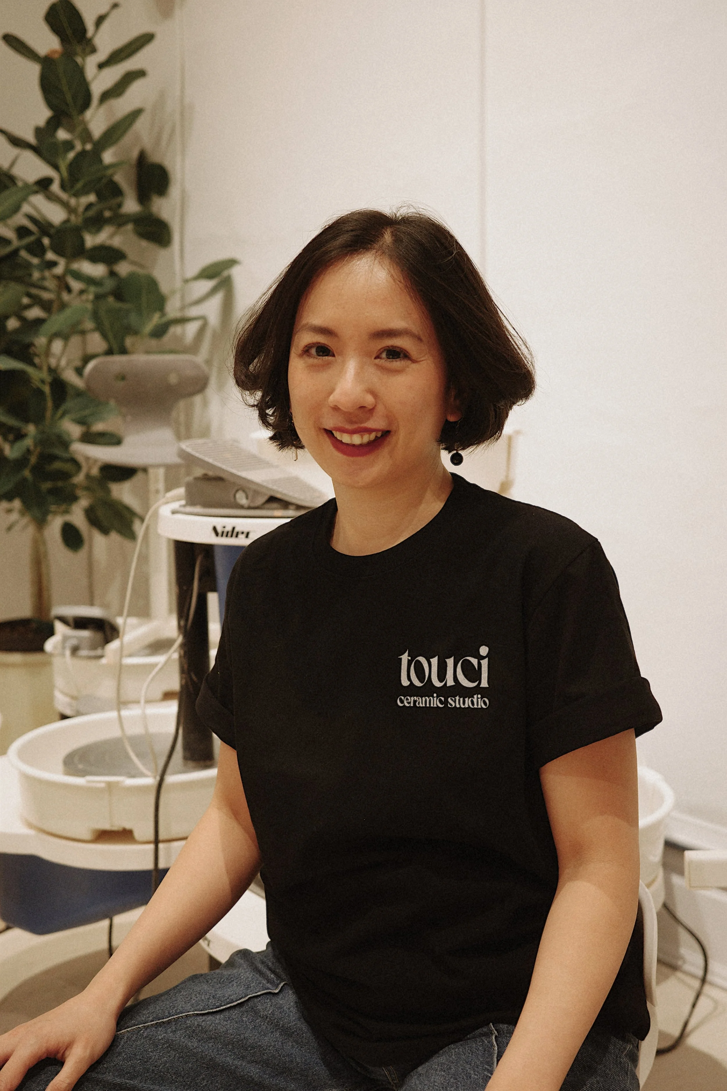 A woman with short brown hair smiling, wearing a black t-shirt with 'touci ceramic studio' logo, sitting in a studio with ceramics equipment and plants in the background.