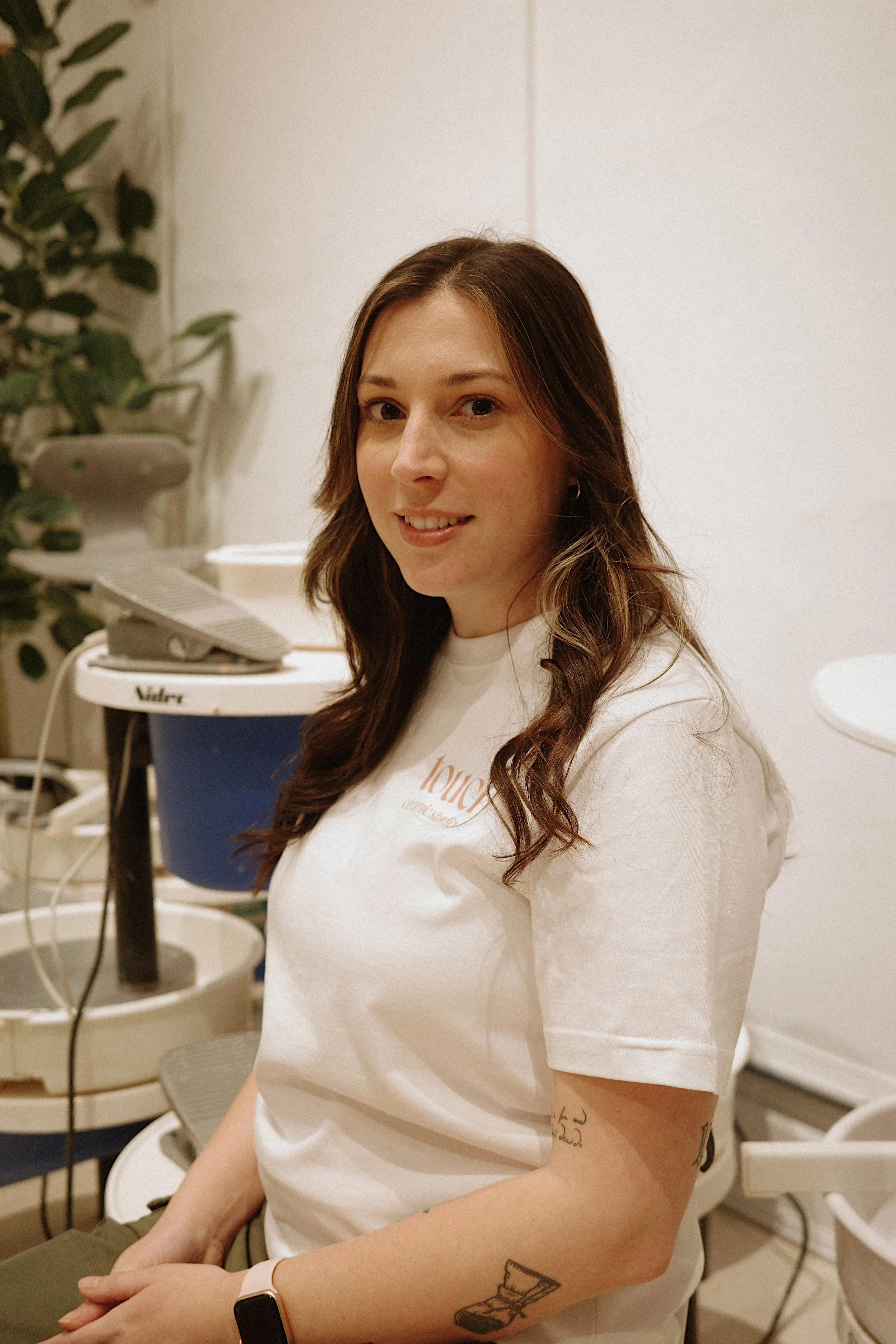 A young woman with light brown, wavy hair, wearing a white t-shirt, seated indoors with medical or cleaning equipment in the background.