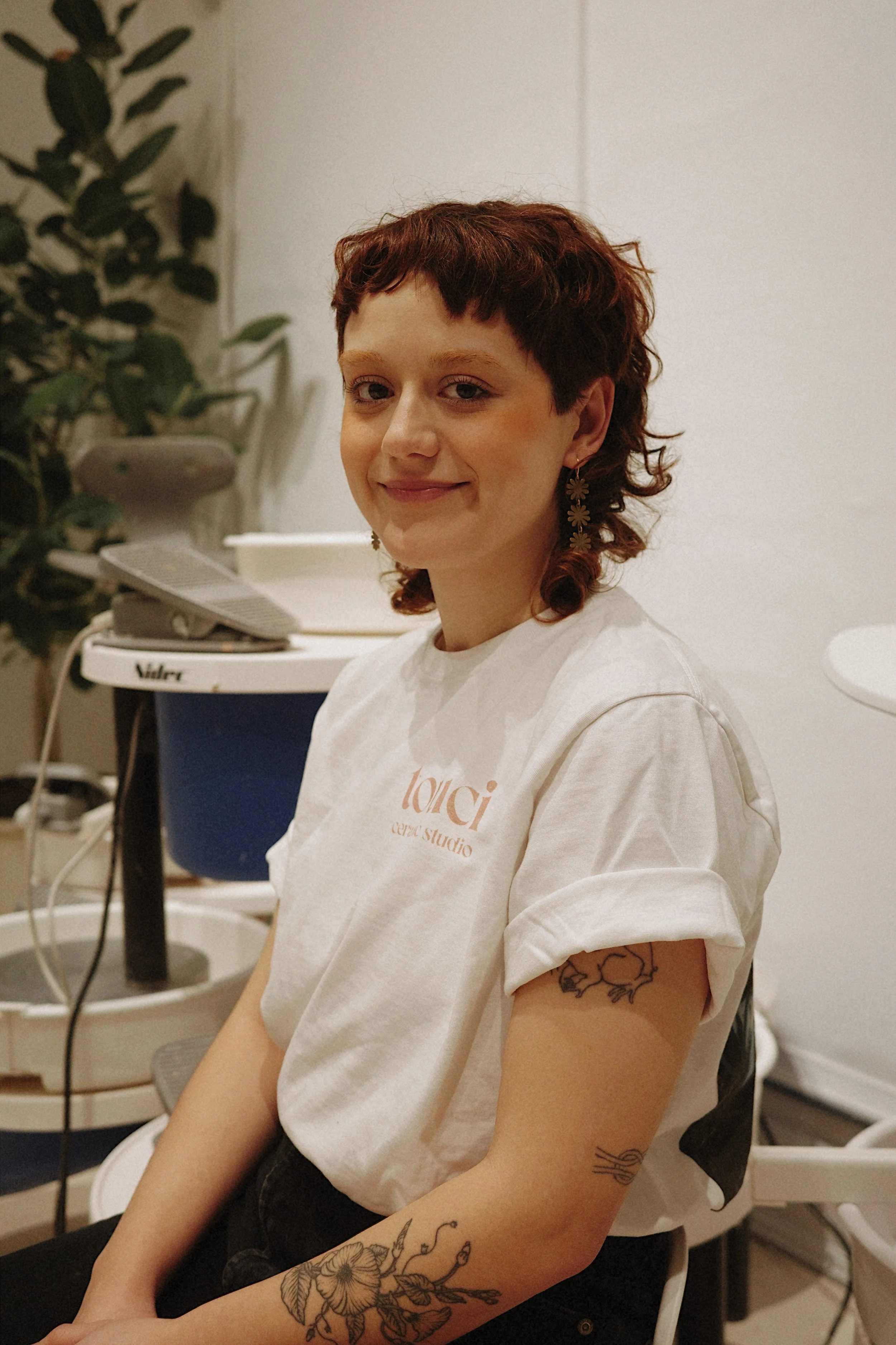 A young woman with short, curly red hair and tattoos on her right arm, wearing a white t-shirt with the logo 'loui ceres studio'. She is smiling and sitting in a room with a plant and some electronic equipment in the background.