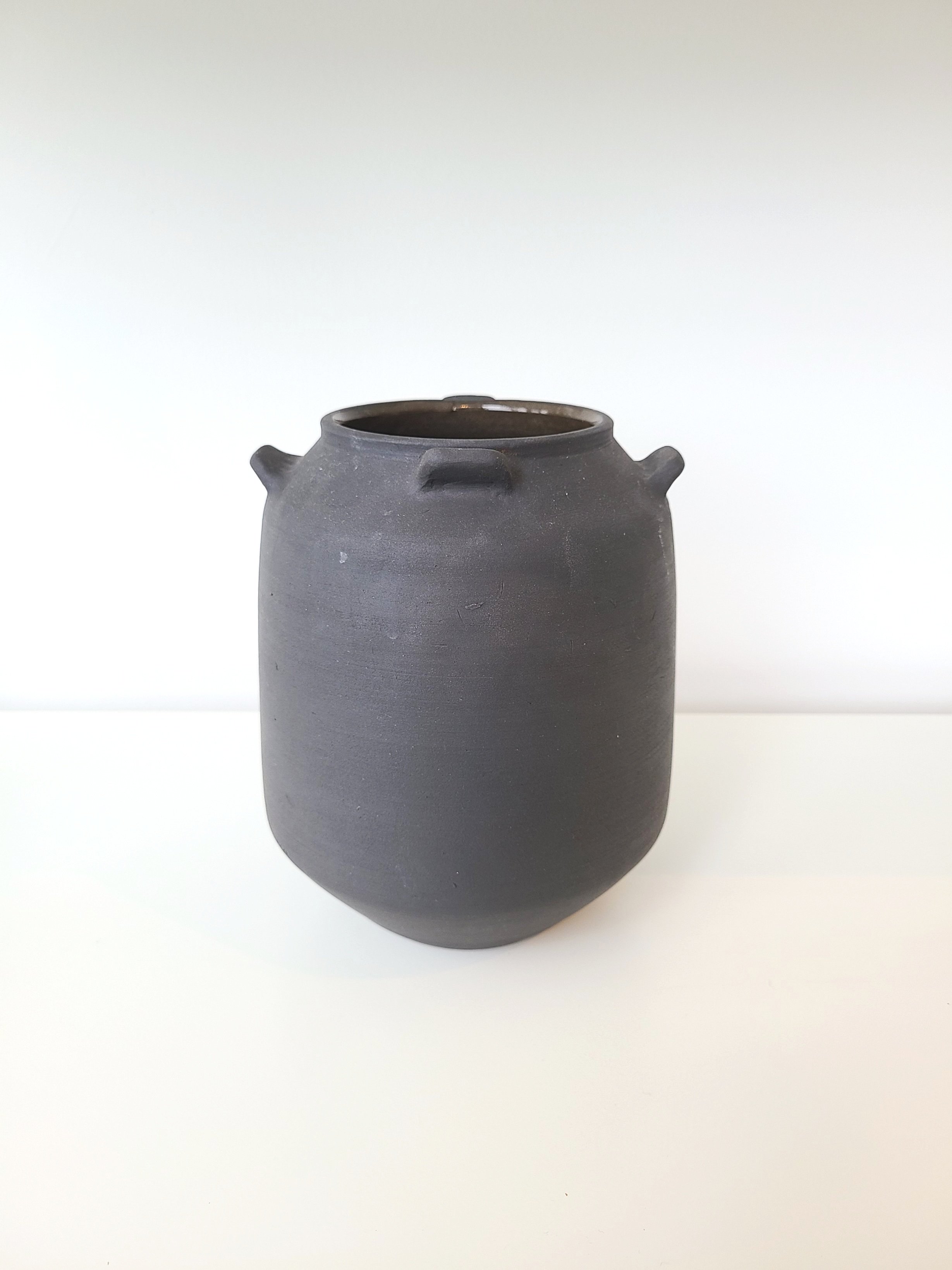 A black ceramic jar with a rounded body and small handles, placed on a white surface against a plain white background.