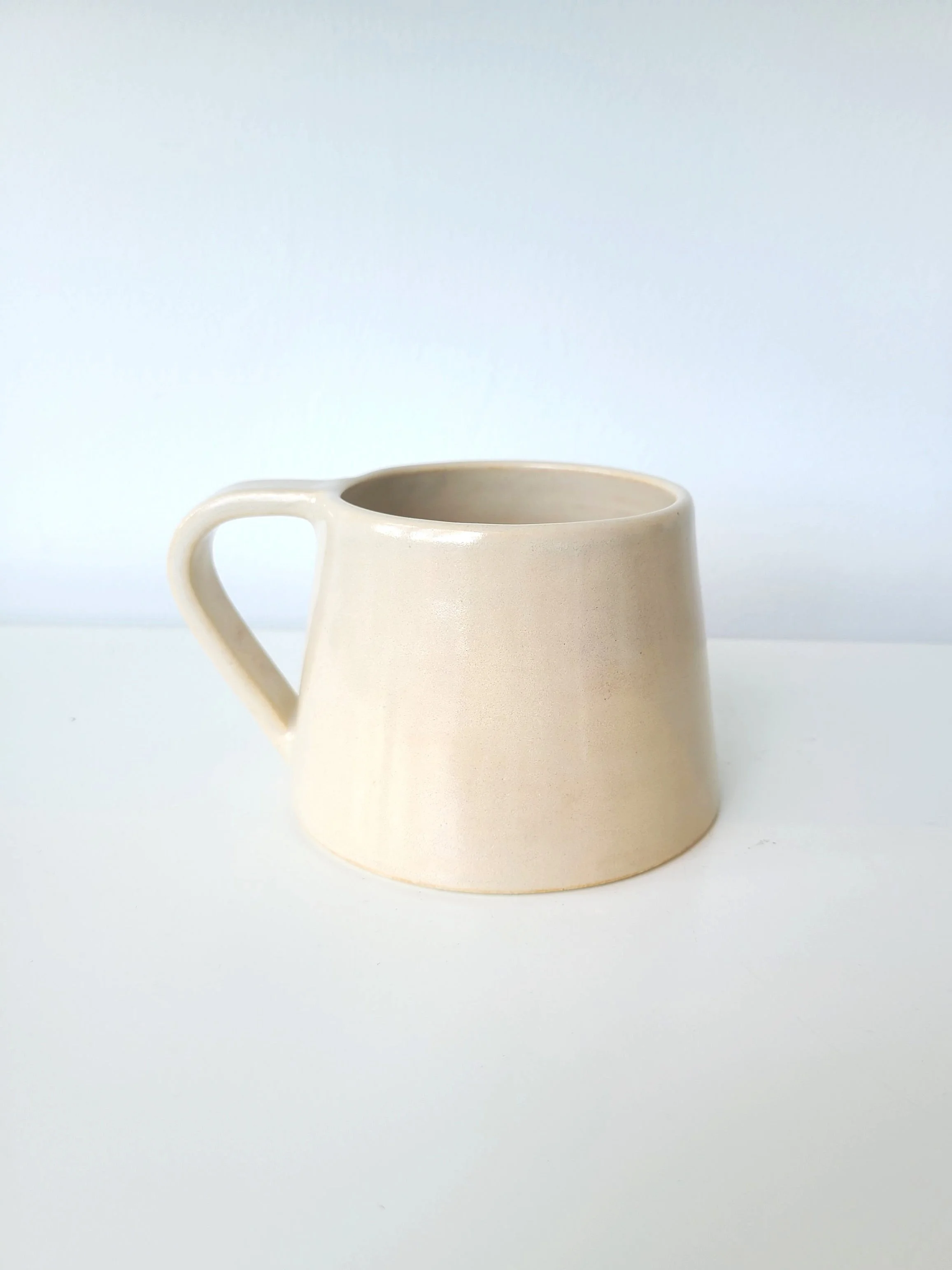 A beige ceramic mug with a handle, placed on a white surface with a light background.
