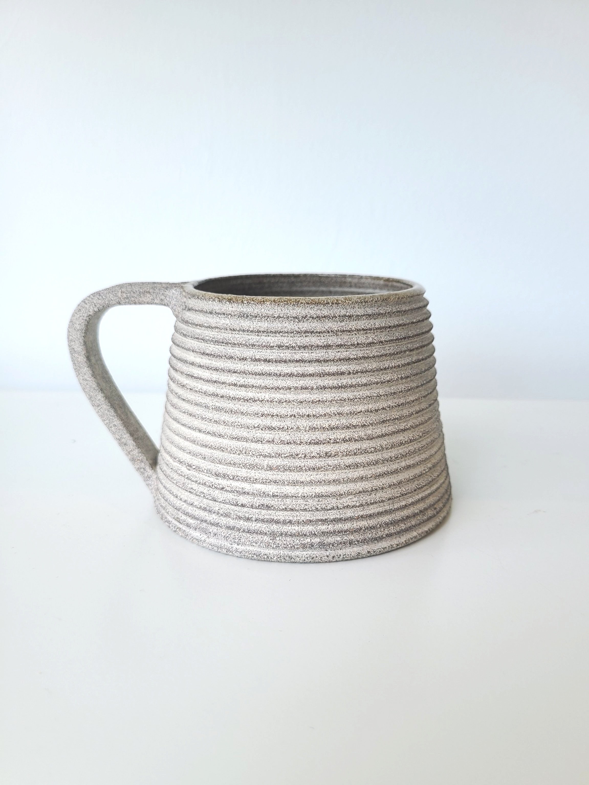 A textured, grey ceramic mug with horizontal ridges and a handle, placed on a white surface with a plain white background.