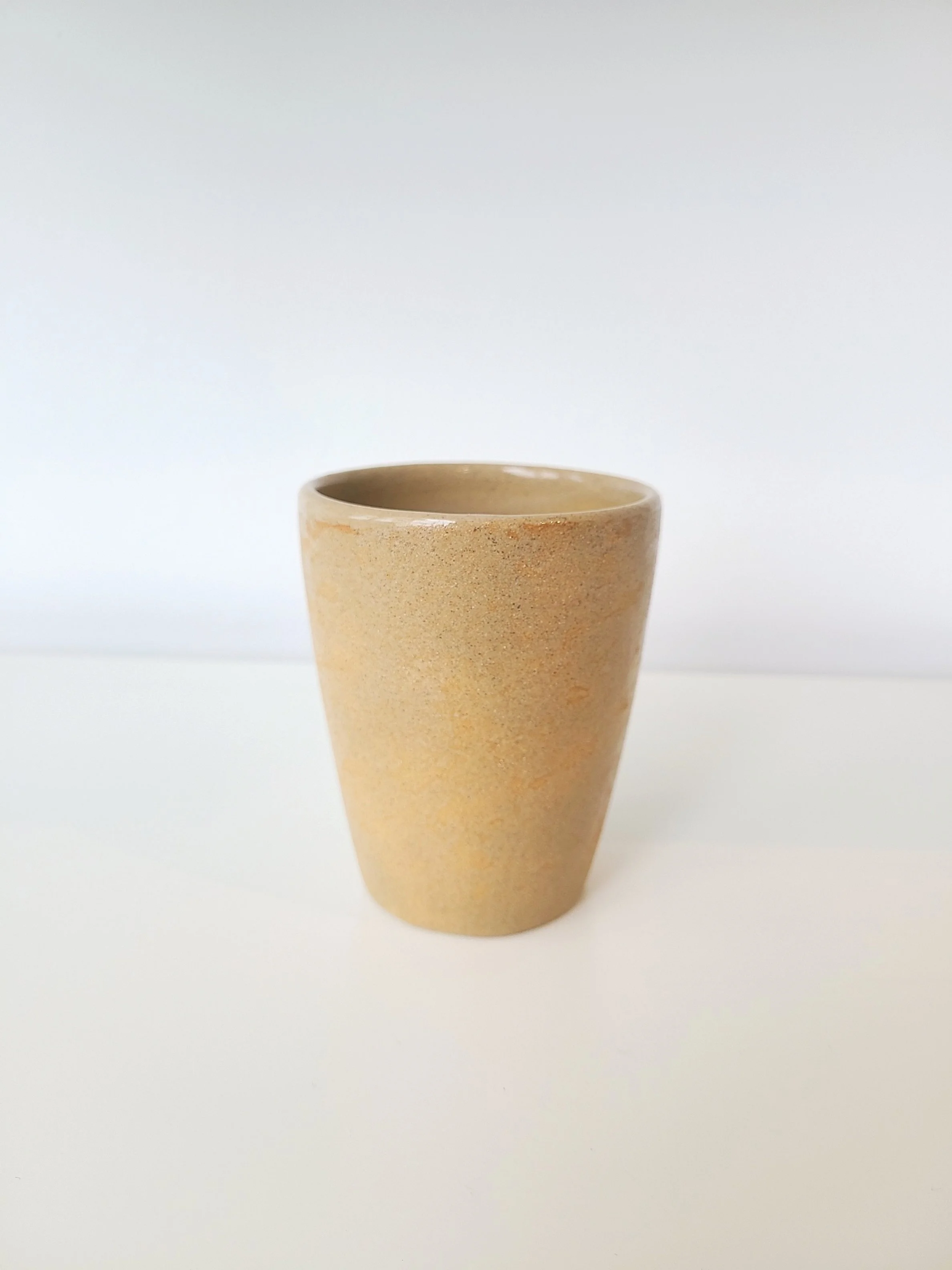 A beige ceramic cup with a smooth surface placed on a white surface against a white background.