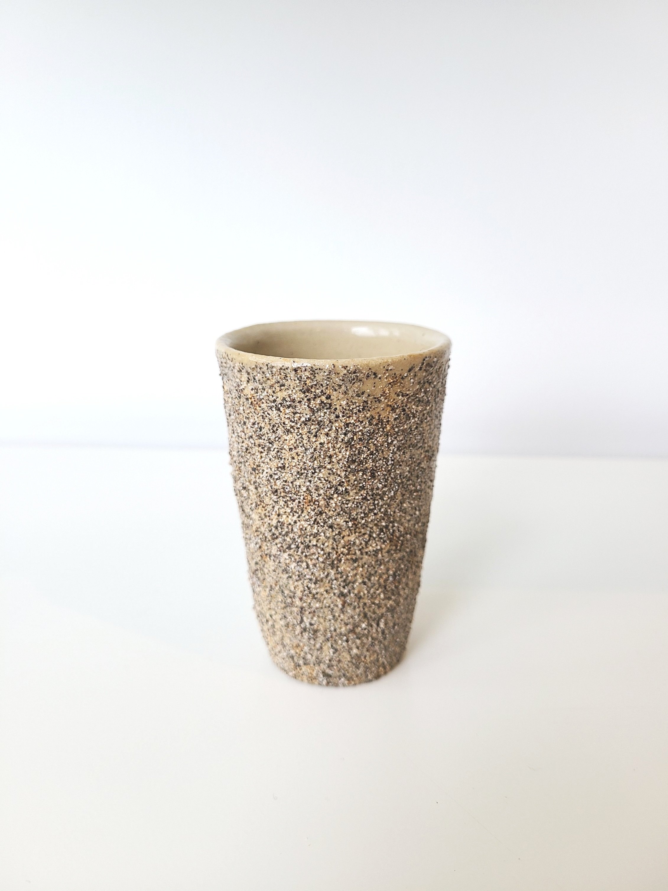A speckled ceramic cup or tumbler with a rough textured exterior and a smooth interior, placed on a white surface against a white background.